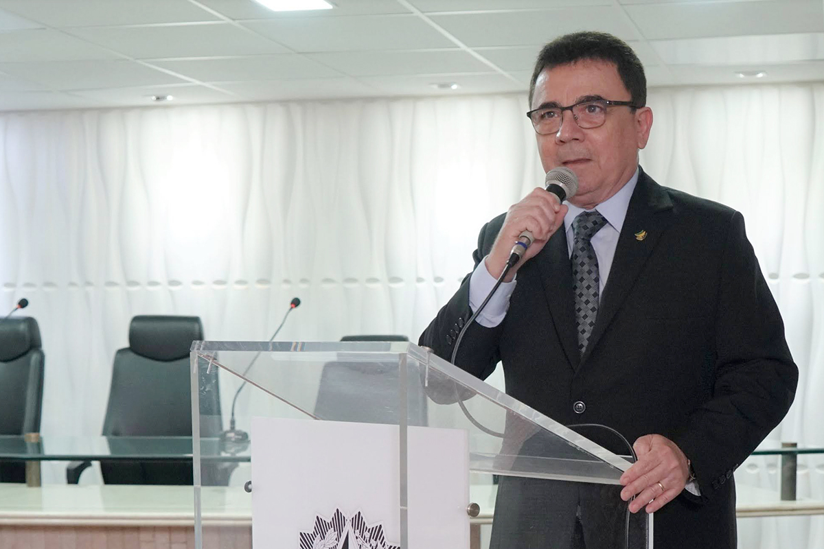 Em plano médio, um homem de pele clara, cabelos curtos e óculos fala ao microfone, segurando-o com a mão direita. Ele veste terno preto, camisa branca e gravata estampada em tons de cinza. O orador está posicionado atrás de um púlpito de acrílico transparente. Ao fundo, nota-se uma parede branca com revestimento em relevo ondulado e encostos de cadeiras pretas.