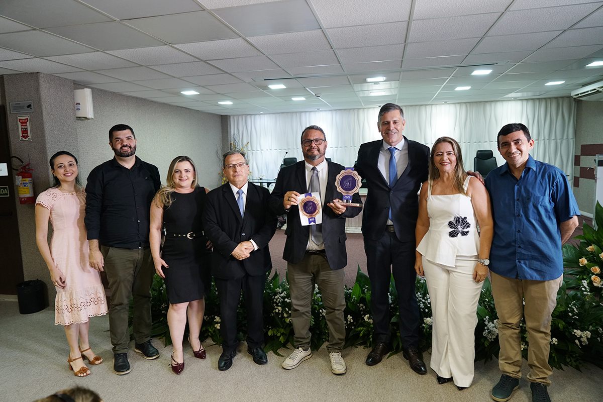 Sete pessoas posam para foto em pé, alinhadas lado a lado em um salão com carpete. O homem ao centro, de óculos, barba e paletó cinza, segura um troféu do "Selo Excelência" e um certificado, ladeado por três homens e três mulheres sorridentes. O grupo veste roupas variadas, incluindo camisas sociais, vestidos e calças jeans. O ambiente possui teto rebaixado com iluminação branca.