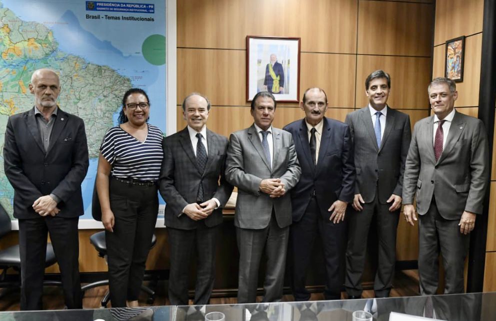 Foto oficial de sete pessoas posando de pé em um gabinete. O grupo, composto por seis homens em trajes formais e uma mulher de blusa listrada, está alinhado à frente de uma mesa de madeira. Na parede de fundo, aparecem um mapa do Brasil e o retrato oficial do Presidente da República. O clima é de cordialidade e parceria institucional.