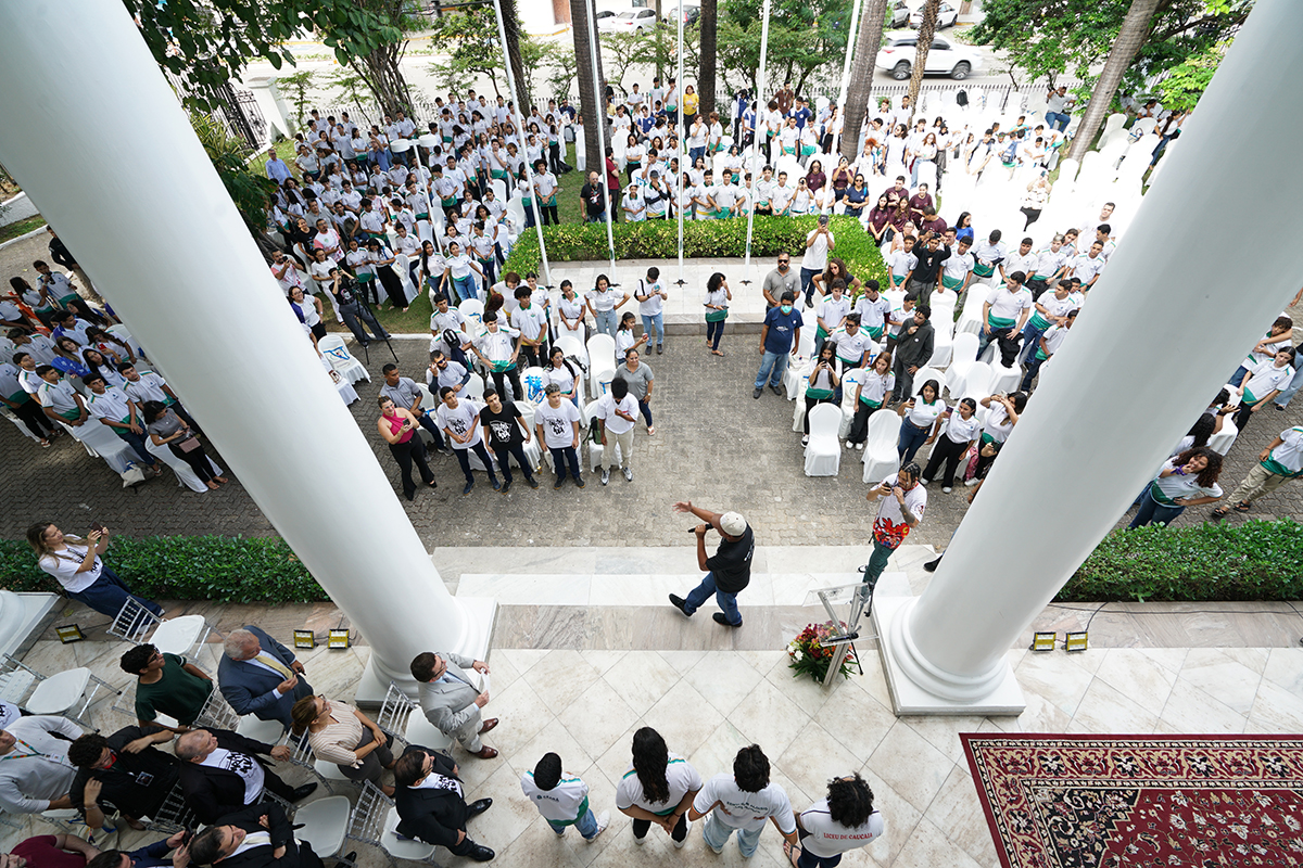 Uma vista aérea de um grande grupo de pessoas reunidas em um pátio externo de mármore e grama. A maioria das pessoas veste camisetas brancas ou a camisa polo com listras verdes e amarelas. Um homem no centro está dançando (provavelmente fazendo um movimento de breakdance), com os braços abertos e boné. Quatro grandes colunas brancas verticais emolduram a cena, com o público espalhado ao redor e nas áreas verdes.