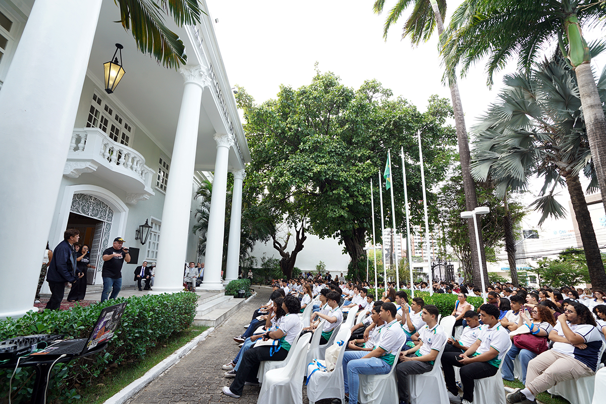 Uma grande audiência de jovens, muitos vestindo a mesma camisa polo branca com listras verdes e amarelas, está sentada em cadeiras brancas ao ar livre. Eles estão em frente a um grande prédio branco com colunas e varandas. No canto esquerdo, ao lado do prédio, algumas pessoas, incluindo um homem de boné preto, estão de pé. A cena é em um gramado cercado por árvores altas e palmeiras, sob um céu claro.
