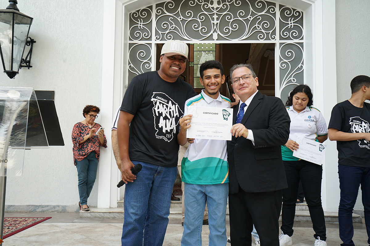Três homens sorriem em frente à entrada decorada de um prédio branco. O da esquerda, moreno e de boné, veste uma camiseta preta estampada e calça jeans, segurando um microfone na mão. O do centro, um jovem em uma camisa polo branca com listras verdes e amarelas, segura um certificado. À direita, um homem branco de terno preto e gravata azul posa ao lado do jovem. Atrás, outras pessoas, incluindo uma mulher em polo branca.