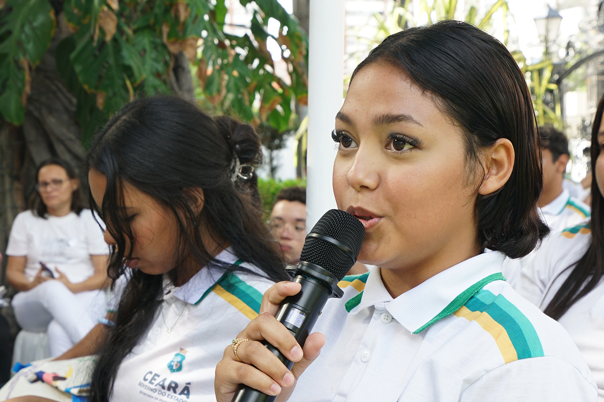 m close-up de uma jovem de pele morena clara falando em um microfone preto, segurando-o com a mão. Ela tem cabelo escuro na altura dos ombros e veste a camisa polo branca com listras verdes e amarelas no colarinho e ombro. Ao seu lado, outra jovem está com a cabeça baixa. A imagem foca no rosto da oradora em primeiro plano, com outras pessoas desfocadas ao fundo.