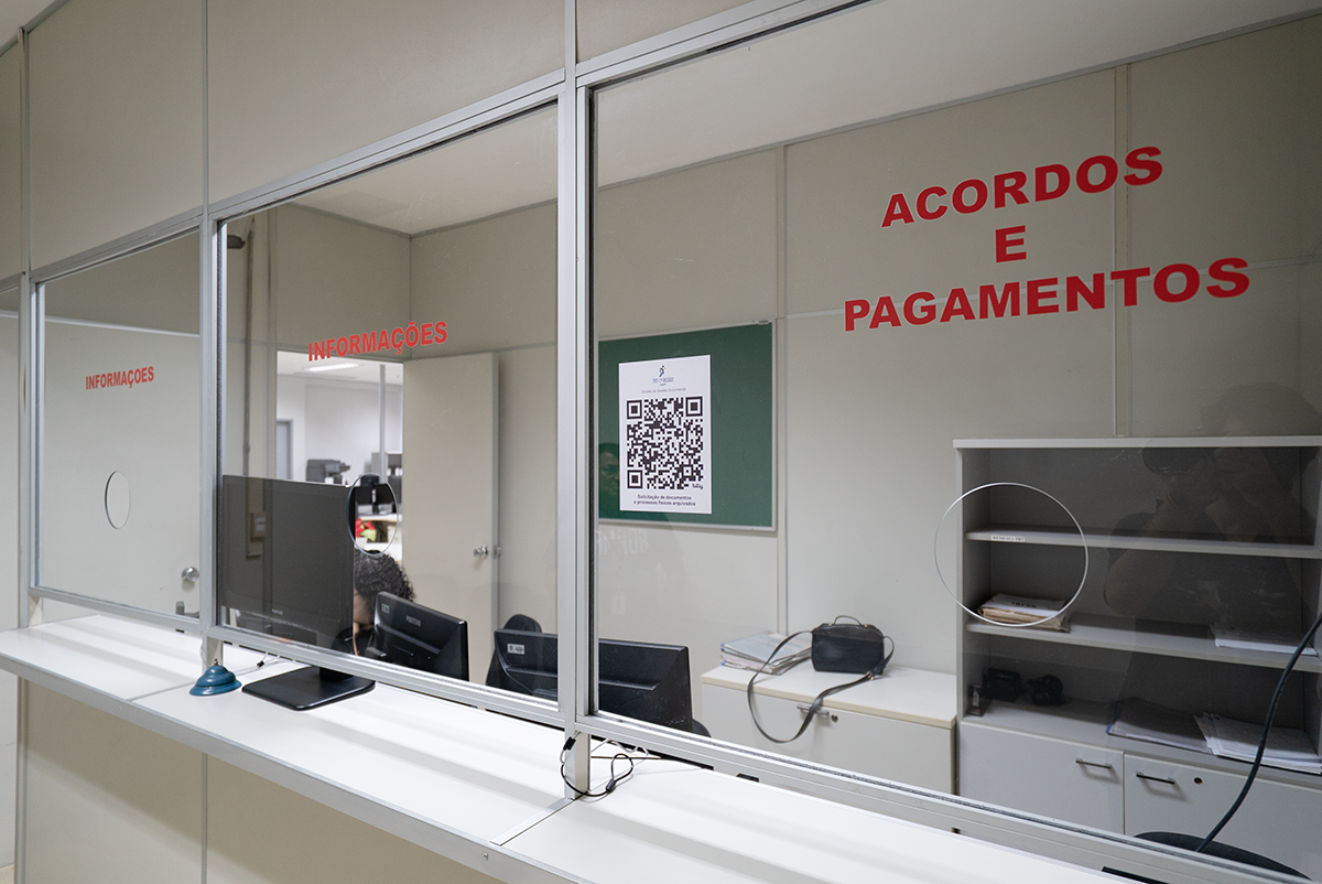 Um balcão de atendimento de vidro com duas divisões. À esquerda, uma placa vermelha diz "INFORMAÇÕES". À direita, outra placa vermelha diz "ACORDOS E PAGAMENTOS". Nesta seção, atrás do vidro, há um quadro verde com um QR Code grande e um pequeno cartaz branco logo acima. Sobre a bancada, vemos monitores de computador e materiais de escritório.