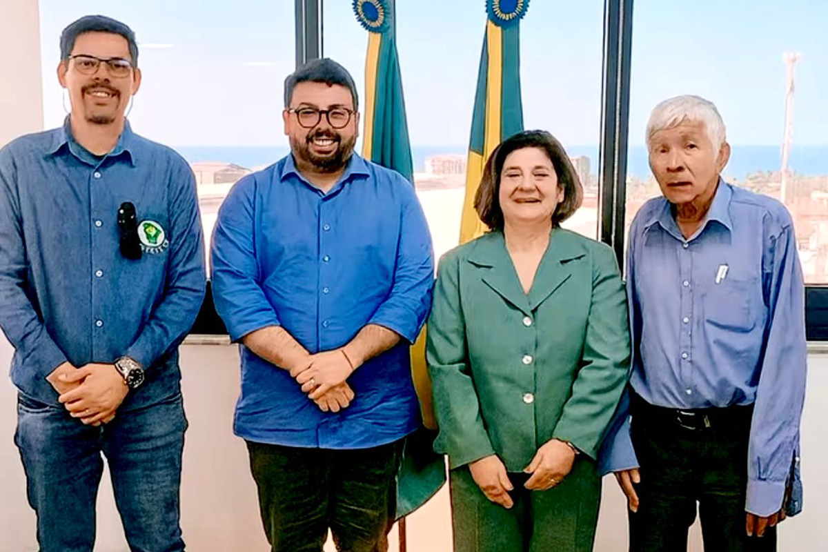 Quatro pessoas estão lado a lado em um ambiente interno. Da esquerda para a direita: um homem de camisa azul, óculos e barba, um homem barbudo de camisa azul-royal, uma mulher de meia-idade sorrindo, vestindo um conjunto de blazer e saia verde musgo, e um senhor mais velho de camisa social azul clara. Atrás deles, há duas bandeiras com detalhes azuis e dourados.
