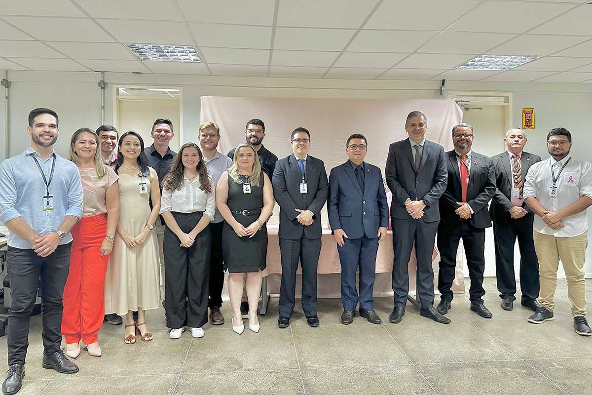 Uma fotografia em plano médio mostra um grupo de quatorze pessoas alinhadas lado a lado em um ambiente de escritório com paredes claras. O grupo, composto por homens e mulheres com trajes casuais e formais, sorri para a câmera à frente de um painel coberto por um tecido rosa claro. Alguns utilizam crachás institucionais e, ao fundo, percebem-se portas e uma luminária de emergência na parede.