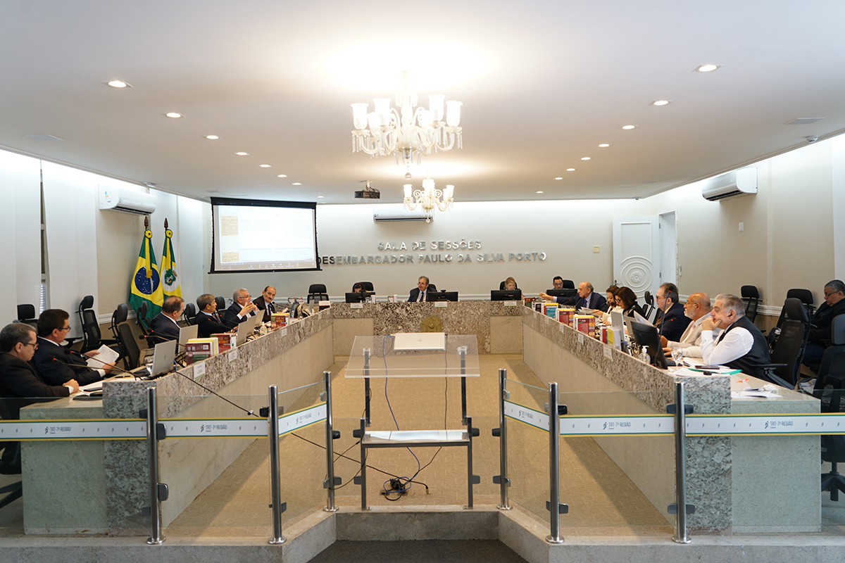 Fotografia de plano aberto da Sala de Sessões do Tribunal Pleno, onde magistrados estão sentados em torno de uma grande bancada em formato de "U". O ambiente formal contém laptops, livros jurídicos e as bandeiras do Brasil e do Ceará ao fundo. Na parede central, destaca-se a inscrição "Sala de Sessões Desembargador Paulo da Silva Porto".