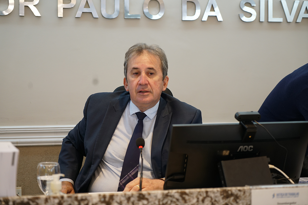 Close-up do desembargador Paulo Régis Machado Botelho, diretor da Escola Judicial. Ele veste terno escuro, camisa clara e gravata azul com detalhes listrados. O magistrado está falando ao microfone, posicionado atrás de um monitor de computador. Ao fundo, parte do letreiro prateado da Sala de Sessões é visível na parede cinza claro.