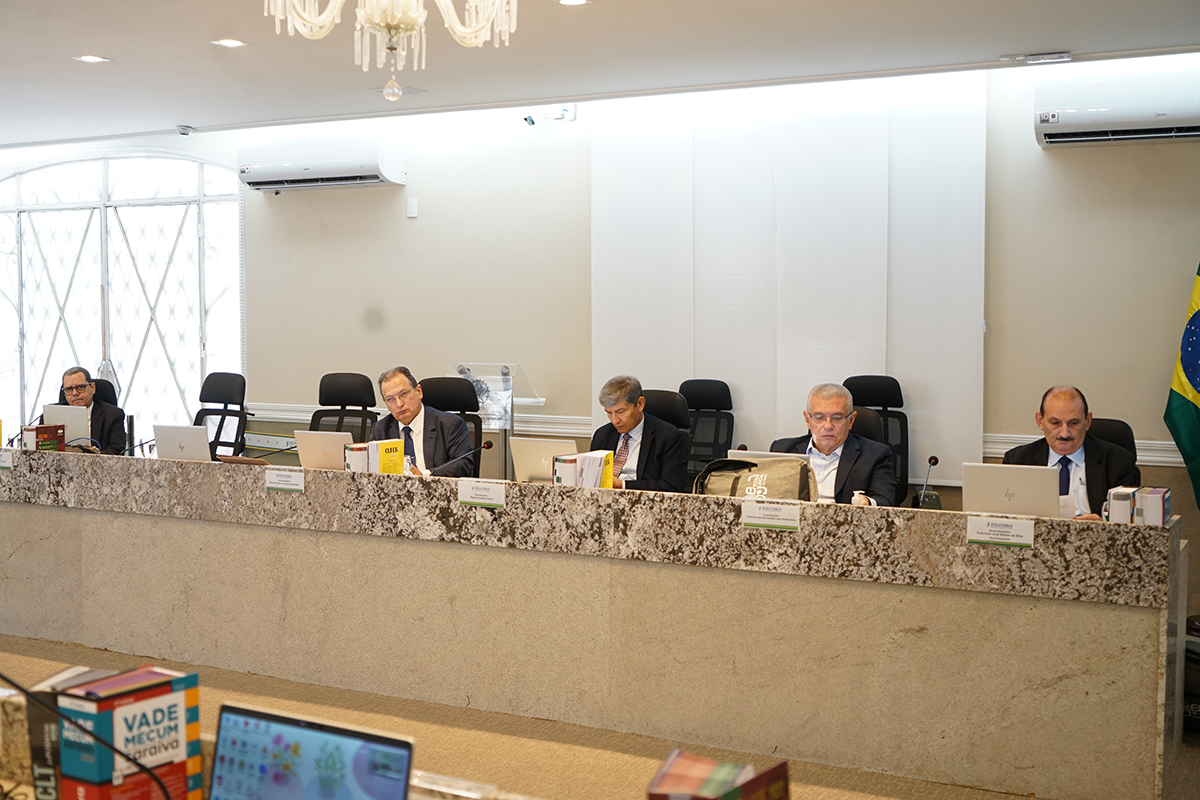 Fotografia em plano médio mostrando cinco desembargadores sentados lado a lado atrás de uma longa bancada de granito salpicado. Eles vestem ternos escuros e estão concentrados em seus laptops e documentos. O ambiente é bem iluminado, com uma grande janela de grades brancas à esquerda e a bandeira do Brasil no canto direito. O desembargador posicionado na extrema direita tem bigode e olha para baixo, analisando papéis