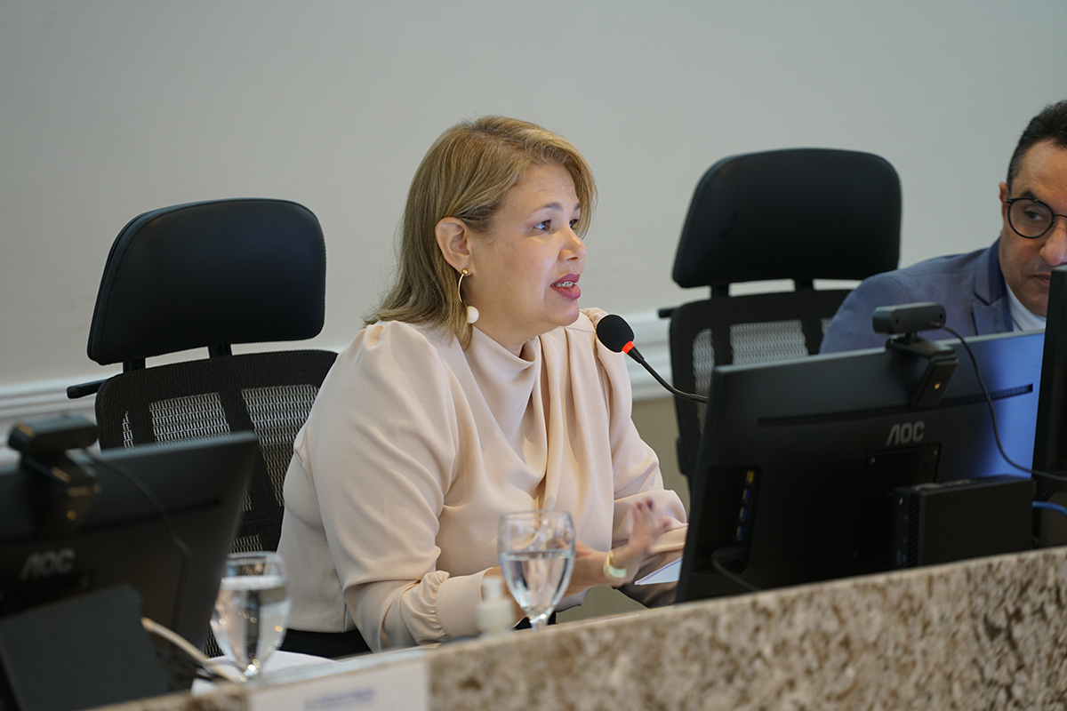 Fotografia em plano aberto da Sala de Sessões do Pleno, iluminada por lustres de cristal. Magistrados estão sentados ao redor de uma grande mesa em formato de "U" de granito, utilizando notebooks e livros jurídicos. Ao fundo, há telas de projeção, as bandeiras do Brasil e do Ceará, e o letreiro da sala