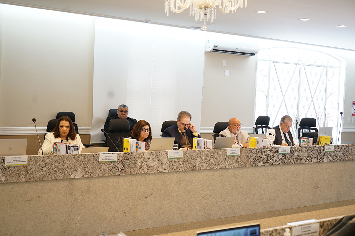 Fotografia em plano aberto da Sala de Sessões Desembargador Paulo da Silva Porto. Os magistrados estão sentados em uma grande mesa de mármore claro em formato de "U", equipada com notebooks e microfones. No centro do teto, destacam-se dois lustres de cristal e, ao fundo, um telão exibe um documento de texto ao lado das bandeiras do Brasil e do Ceará.