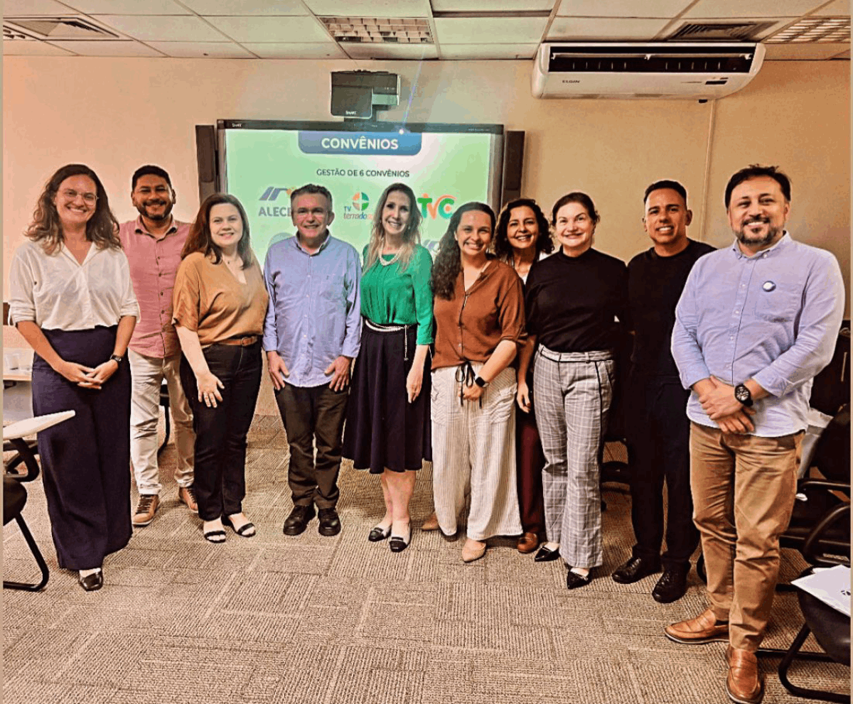 Dez pessoas, sendo seis mulheres e quatro homens, posam sorridentes e alinhadas em uma sala de reuniões com piso acarpetado.  Ao centro, uma mulher de blusa verde e saia midi preta destaca-se diante de uma tela digital que exibe o título "CONVÊNIOS" e diversos logotipos.  O grupo veste trajes sociais e casuais finos, compondo um cenário de integração profissional em um ambiente institucional iluminado.