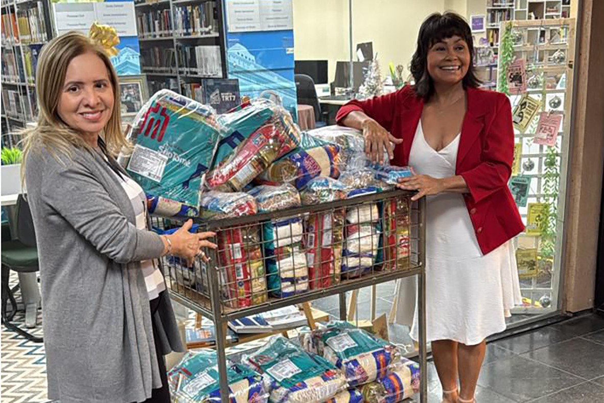 Duas mulheres sorridentes, de meia-idade, posam ao lado de um carrinho metálico cheio de sacos de alimentos embalados, destinados a doações. O ambiente é interno e iluminado, com prateleiras de biblioteca repletas de livros e decorações de Natal ao fundo. A mulher à direita veste um blazer vermelho e um vestido branco; a outra, um casaco cinza claro.