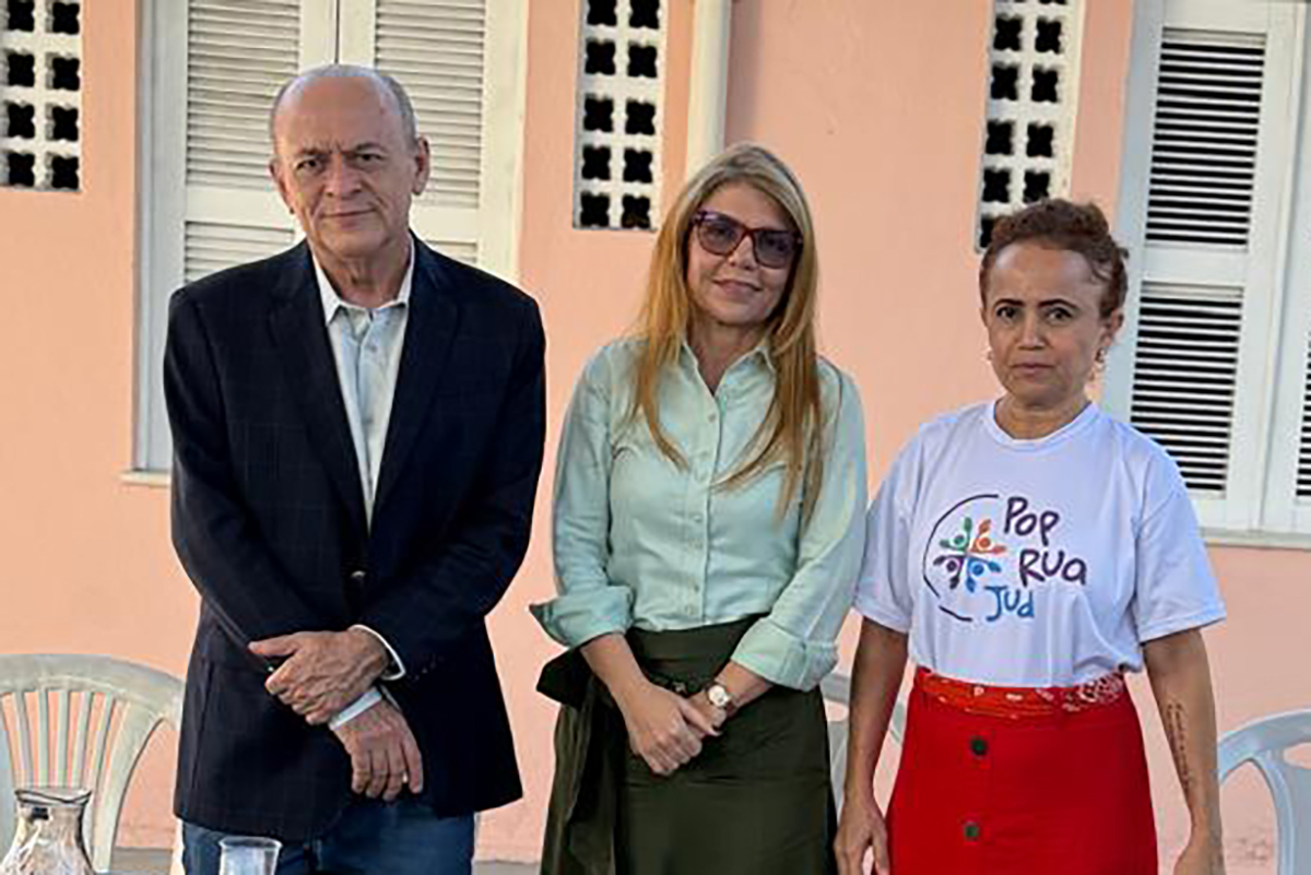 Foto de três pessoas em pé (um homem e duas mulheres), pousada e estão na frente de uma casa de parede marfim e janelas brancas.