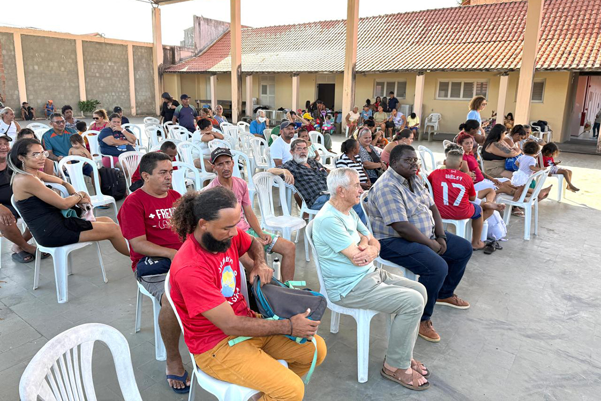 Uma assembleia em área externa durante o dia: um grande grupo de pessoas, de diversas idades, senta-se em cadeiras de plástico brancas. O público, em sua maioria, olha para a frente, onde ocorre a atividade. Ao fundo, vê-se um muro alto e construções com telhados.
