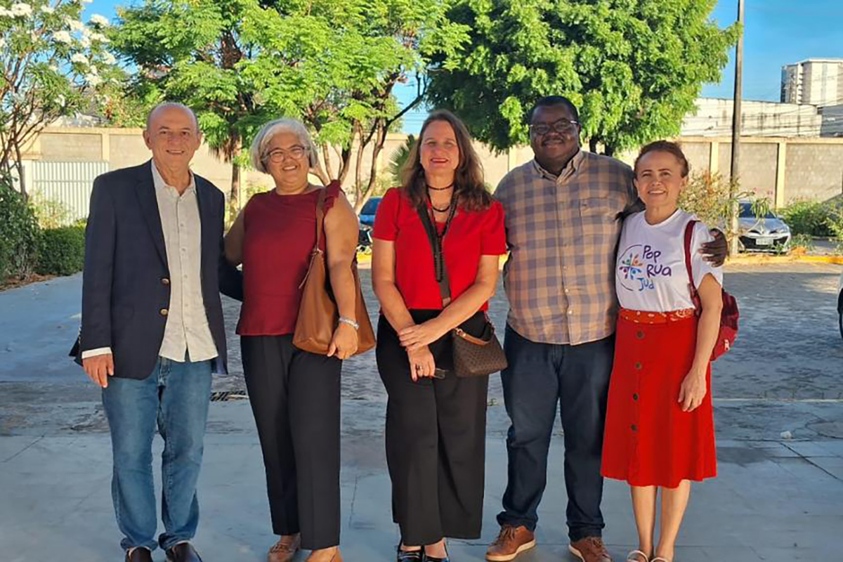 Cinco pessoas — duas mulheres e três homens — estão lado a lado, sorrindo para a foto. A pessoa na extremidade direita veste uma camiseta branca com a estampa "Pop Rua Jud". Eles estão em um ambiente externo, com árvores verdes frondosas ao fundo e o chão claro.