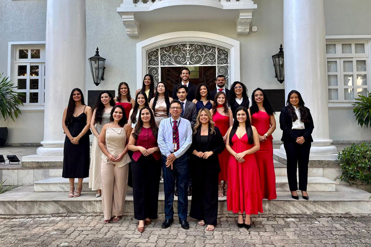  Uma foto em grupo reúne 20 pessoas sorridentes nos degraus de uma imponente construção branca com grandes colunas. À frente, um homem de camisa social azul e gravata vermelha está ladeado por mulheres em trajes formais (tons de bege, vinho, preto e vermelho). Atrás deles, os demais integrantes se distribuem em fileiras, vestindo roupas elegantes em uma paleta predominante de vermelho, preto e branco. O ambiente é externo, durante o dia, sugerindo uma cerimônia ou evento oficial.