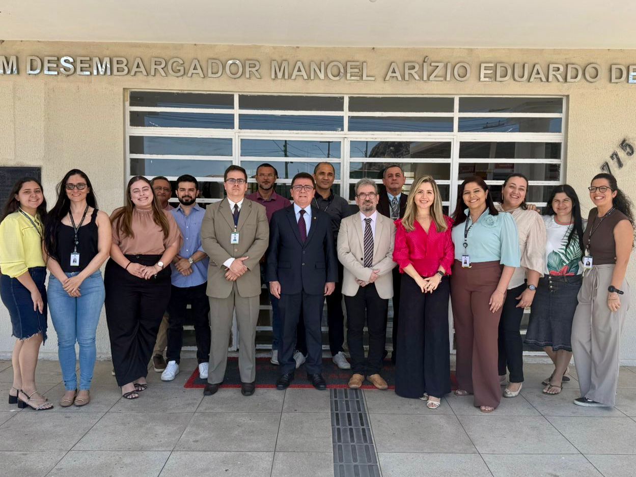A foto mostra um grupo de dezesseis pessoas, composto por homens e mulheres, posando em pé, lado a lado, em frente à fachada de um prédio público.  As pessoas estão sorrindo e olhando para a câmera. No centro do grupo, destacam-se homens vestidos formalmente com terno e gravata, enquanto os demais usam roupas sociais ou esporte fino (como camisas, blusas e calças jeans/sociais); algumas pessoas portam crachás funcionais no pescoço.  Ao fundo, a parede do prédio é de cor creme e possui uma grande estrutura de vidro com grades brancas horizontais. Acima das cabeças do grupo, há um letreiro prateado fixado na parede onde se lê parcialmente: “DESEMBARGADOR MANOEL ARÍZIO EDUARDO DE”. O chão é calçado com pedras claras e a iluminação indica um dia ensolarado.
