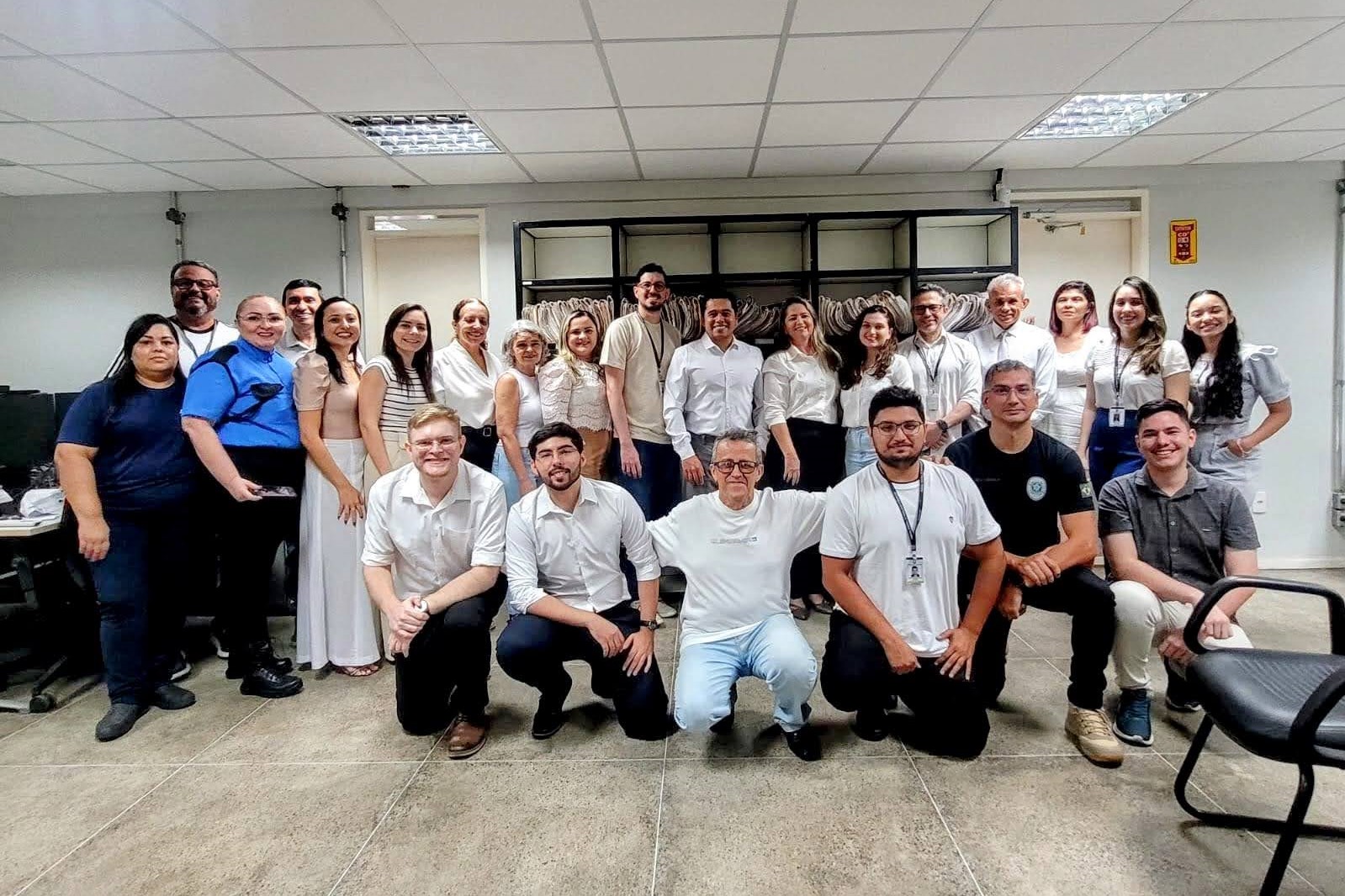 Fotografia colorida horizontal em plano inteiro de um grupo grande em um escritório.  Cenário: O ambiente é um escritório bem iluminado com piso de cerâmica cinza e teto branco com luminárias fluorescentes. Ao fundo, centralizada, há uma grande estante metálica preta preenchida com pastas de arquivos suspensos.  Personagens: O grupo é composto por cerca de 27 pessoas, homens e mulheres adultos de diversas idades. Todos olham para a câmera e sorriem.  Disposição: As pessoas estão organizadas em dois níveis. A grande maioria forma uma linha em pé que ocupa toda a largura da foto. À frente deles, seis homens estão agachados ou ajoelhados.  Vestuário: A maioria veste roupas no estilo esporte-fino ou casual de trabalho, com predominância de camisas brancas ou tons claros. Muitos utilizam crachás pendurados no pescoço. Destacam-se uma mulher à esquerda usando uma camisa azul-royal vibrante e um homem agachado à direita vestindo uma camiseta preta com um emblema (tipo brasão) no peito.
