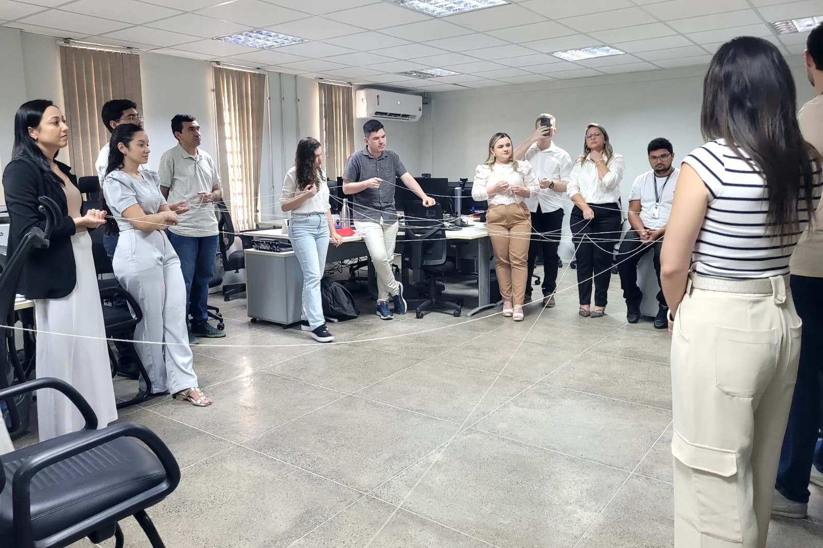 Fotografia colorida horizontal de uma dinâmica de grupo em um escritório.  Cenário: O ambiente é uma sala corporativa ampla, com piso de cerâmica cinza, paredes brancas e janelas ao fundo cobertas por persianas verticais bege. Atrás do grupo, veem-se estações de trabalho com computadores, monitores e cadeiras de escritório pretas.  Ação: Um grupo de cerca de 11 pessoas está em pé, formado em um círculo. Todos seguram pedaços de um longo barbante branco que se estica de uma pessoa para outra, cruzando o centro do círculo várias vezes e formando uma "teia" complexa que conecta todos os participantes.  Personagens: Homens e mulheres adultos vestindo roupas no estilo esporte-fino (camisas sociais, polos e calças de sarja ou jeans).  Em destaque no primeiro plano, à direita, vê-se uma mulher de costas para a câmera vestindo uma blusa listrada e calça bege.  Ao fundo, um homem levanta um celular para tirar uma foto ou filmar a atividade.  Atmosfera: A cena sugere um exercício de integração de equipe ("team building"), com os participantes atentos à dinâmica da teia.