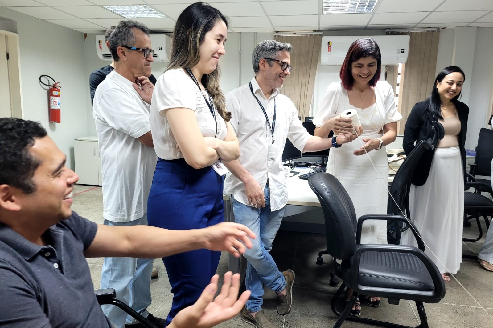 Fotografia colorida horizontal em plano médio de participantes interagindo.  Ação: O foco está em um subgrupo de seis pessoas que sorriem e observam a condução da dinâmica. Uma mulher de vestido branco, ao centro-direita, segura o rolo de barbante branco, preparando-se para passá-lo.  Personagens:  No canto inferior esquerdo, um homem de camisa polo escura sorri e gesticula com as mãos abertas.  Ao centro, um homem de camisa branca e óculos observa a ação com um sorriso discreto, enquanto uma mulher de blusa branca e calça azul está com os braços cruzados ao seu lado.  À direita, uma mulher de blazer preto e saia branca observa a cena alegremente.  Cenário: Parte do escritório, vendo-se um extintor de incêndio vermelho na parede ao fundo e cadeiras de escritório pretas.