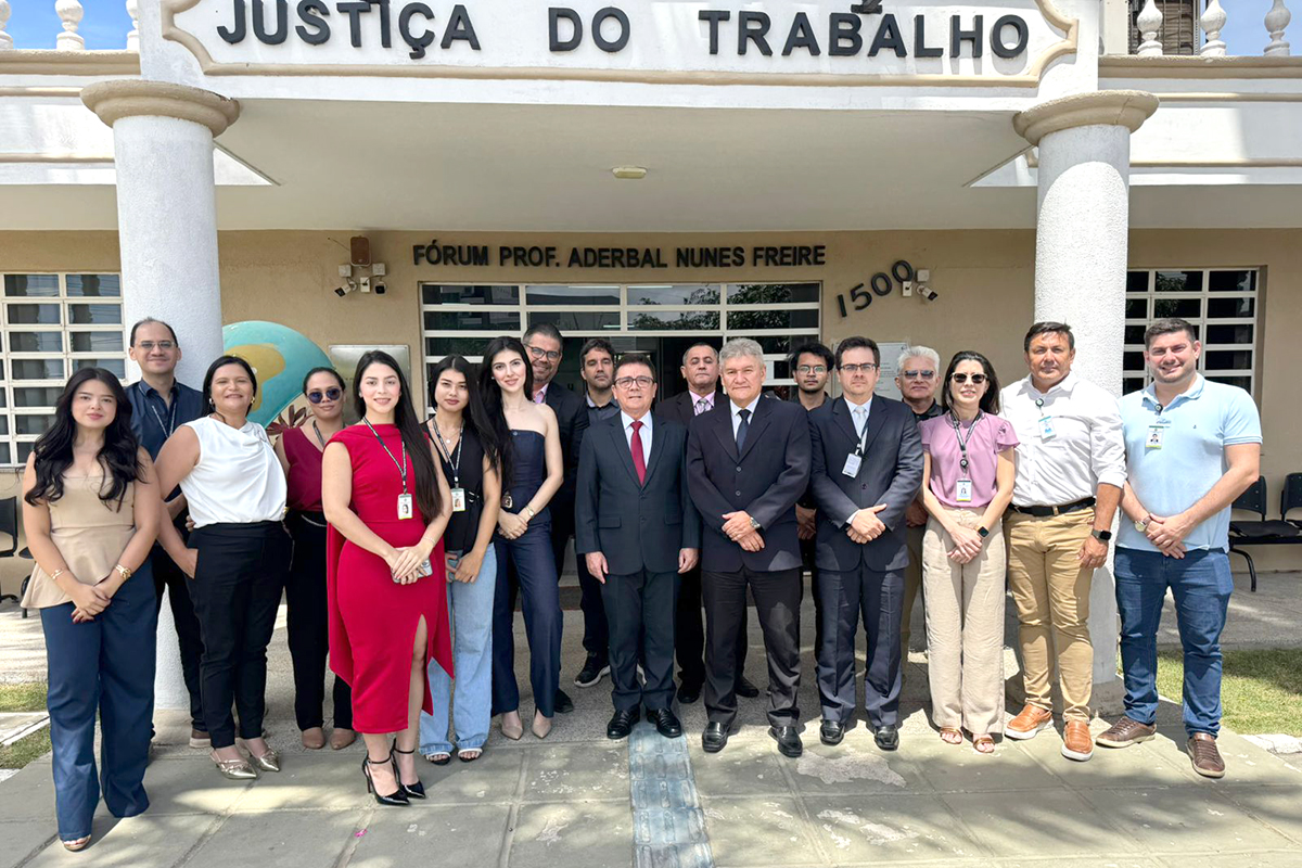 Uma foto em grupo reúne cerca de 18 pessoas em frente ao Fórum Prof. Aderbal Nunes Freire, da Justiça do Trabalho, em Tianguá. Homens e mulheres, com vestimentas que variam entre o social e o casual, posam sorridentes sob a marquise do prédio de tons claros. Ao centro, destacam-se dois homens de terno escuro e uma mulher com um vibrante vestido vermelho. A iluminação é natural e intensa, vinda do sol.