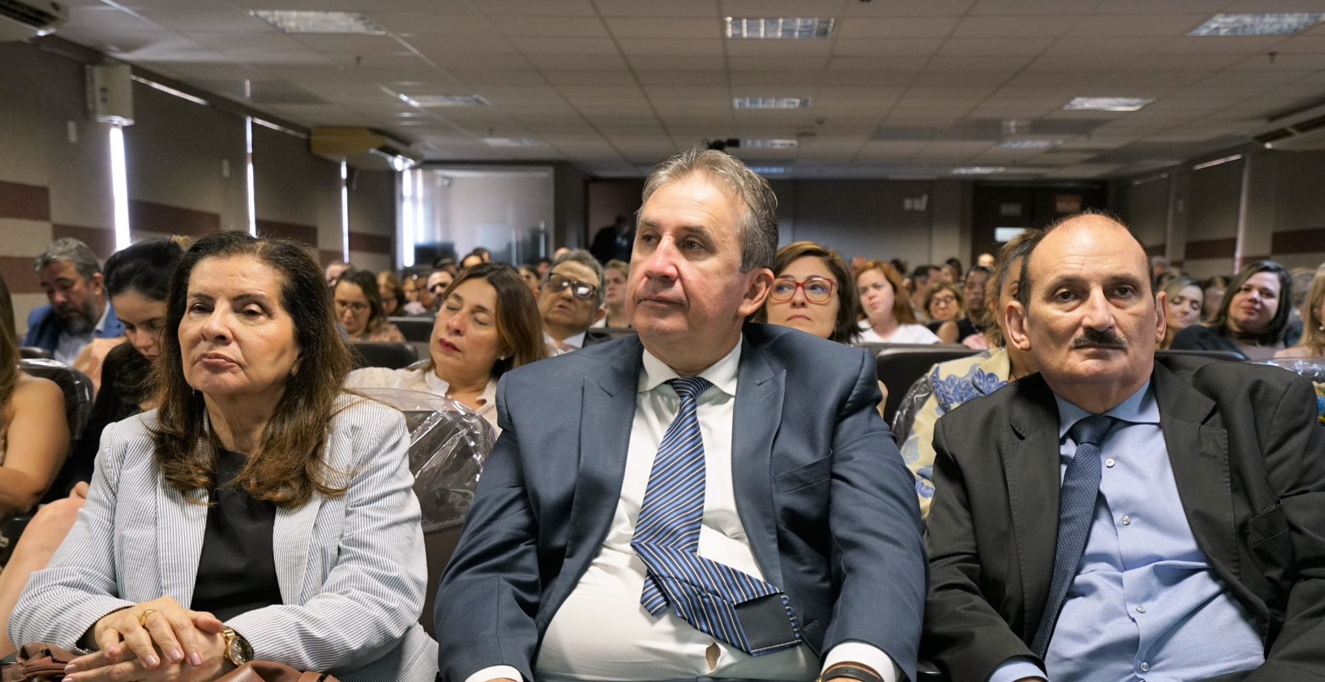 Em um auditório lotado, três pessoas ocupam o primeiro plano: uma mulher à esquerda em um blazer listrado e dois homens de terno e gravata ao centro e à direita. Todos mantêm expressões solenes e atentas, voltadas para o que parece ser o palco do evento. Ao fundo, diversas outras pessoas preenchem as fileiras de poltronas sob a iluminação de um teto com placas de LED.