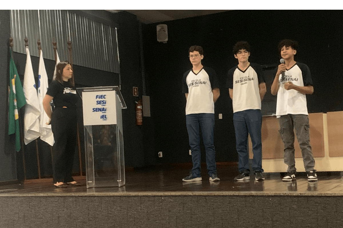 Sobre o palco de um auditório, três estudantes com uniformes brancos e mangas pretas da "Escola SESI SENAI" estão em pé; o jovem à direita fala ao microfone. À esquerda, uma mulher vestida de preto observa os alunos próxima a um púlpito de acrílico e bandeiras oficiais. O fundo é composto por uma parede preta e um painel de metal ondulado, sob iluminação direta.