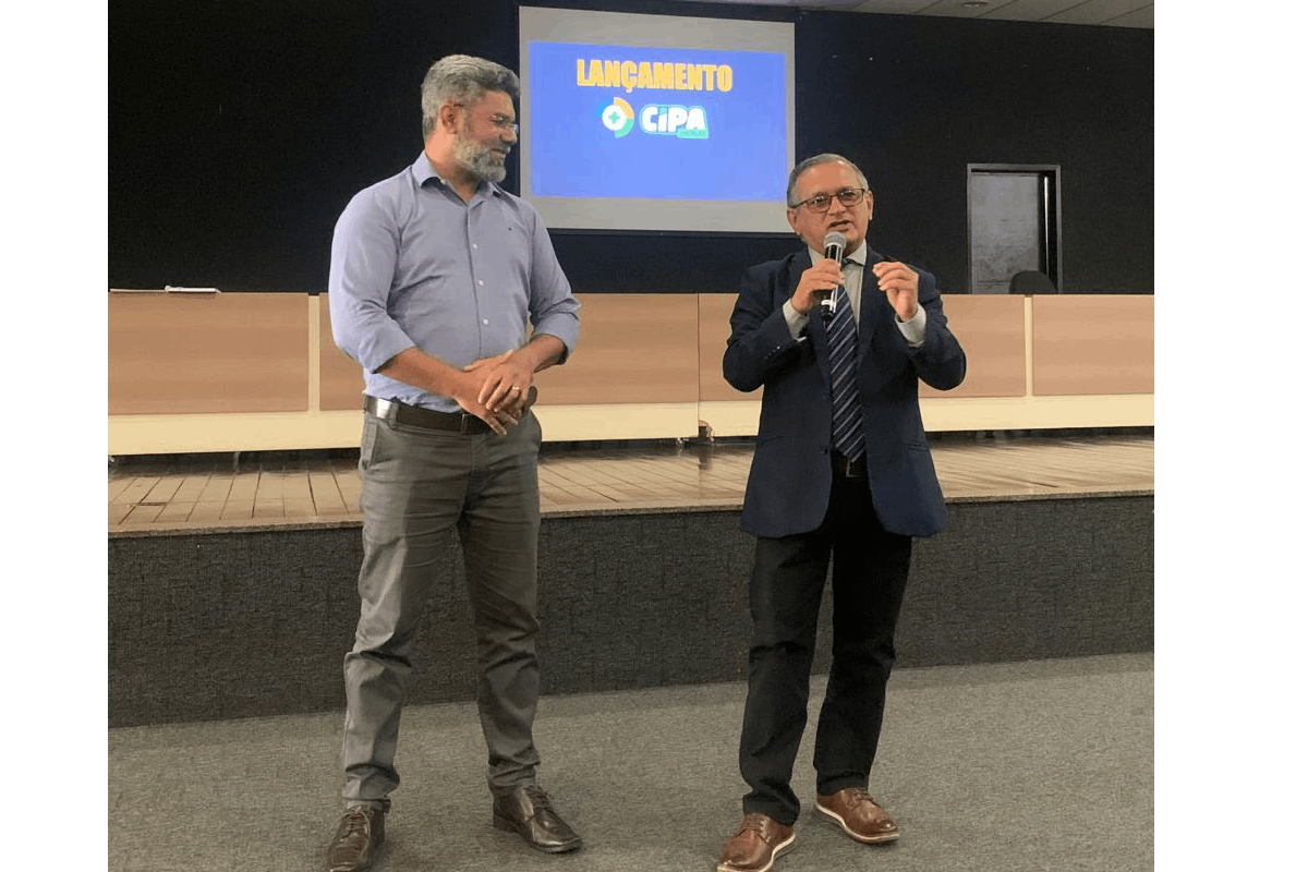 Três estudantes com uniformes brancos e mangas pretas da "Escola SESI SENAI" ocupam o centro de um palco; o jovem à direita fala ao microfone. À esquerda, uma mulher de preto observa próxima a um púlpito de acrílico com logos institucionais e bandeiras. O ambiente tem paredes escuras e teto com acabamento metálico industrial.