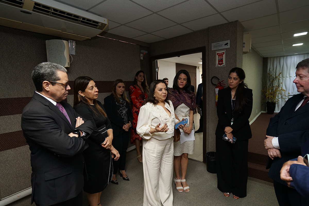 Um grupo de pessoas em trajes formais está reunido em um auditório ou sala de reuniões com paredes revestidas em tecido cinza e marrom. No centro, uma mulher de conjunto branco fala e gesticula, sendo observada atentamente por homens de terno e outras mulheres posicionadas ao seu redor. O ambiente possui iluminação suave e, ao fundo, vê-se uma porta aberta que dá para um corredor mais claro.