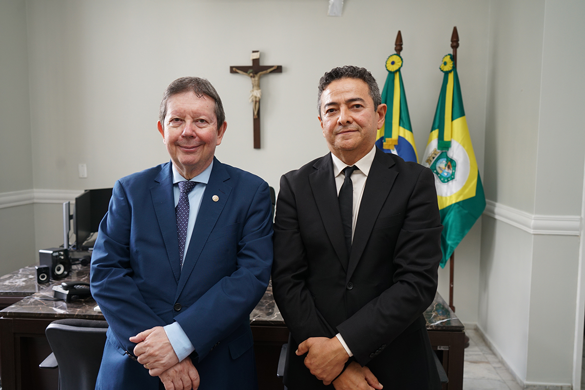 Fotografia colorida, em plano médio e ângulo frontal, mostrando dois homens de pé, lado a lado, em um ambiente de gabinete oficial. À esquerda, um homem de pele clara e cabelos castanhos curtos veste terno azul-marinho, camisa azul-clara e gravata azul-celeste. À direita, um homem de pele morena e cabelos pretos curtos usa terno preto, camisa branca e gravata preta. Ambos estão com as mãos cruzadas à frente do corpo e olham serenamente para a câmera.  Ao fundo, entre os dois homens, há um crucifixo de madeira fixado na parede clara. À direita, aparecem as bandeiras do Brasil e do Estado do Ceará em mastros metálicos. À frente deles, nota-se parte de uma mesa de escritório escura com um monitor de computador à esquerda e pilhas de documentos à direita.
