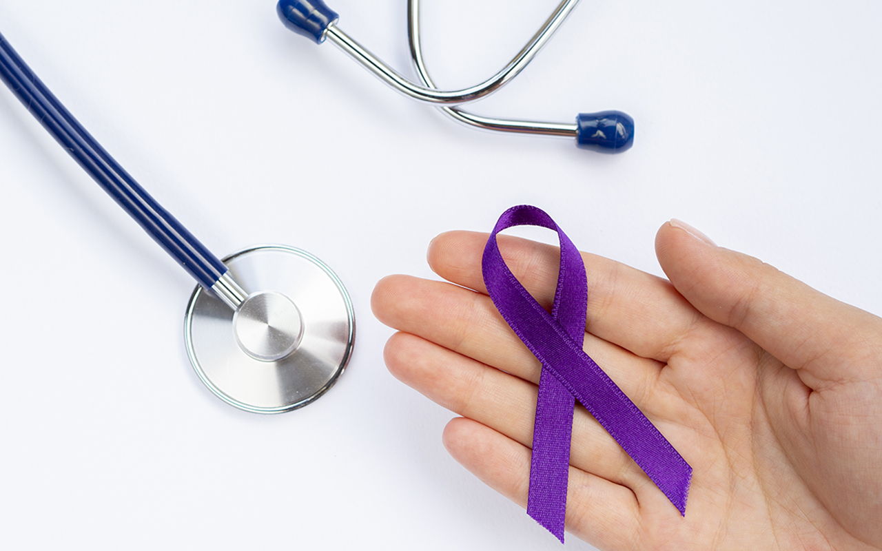 Sobre um fundo branco liso, um estetoscópio azul com fone e campânula prateada está posicionado à esquerda. No centro, uma mão aberta segura um laço de fita roxa, símbolo da campanha Fevereiro Roxo. A imagem transmite uma mensagem de cuidado médico e conscientização sobre doenças crônicas.