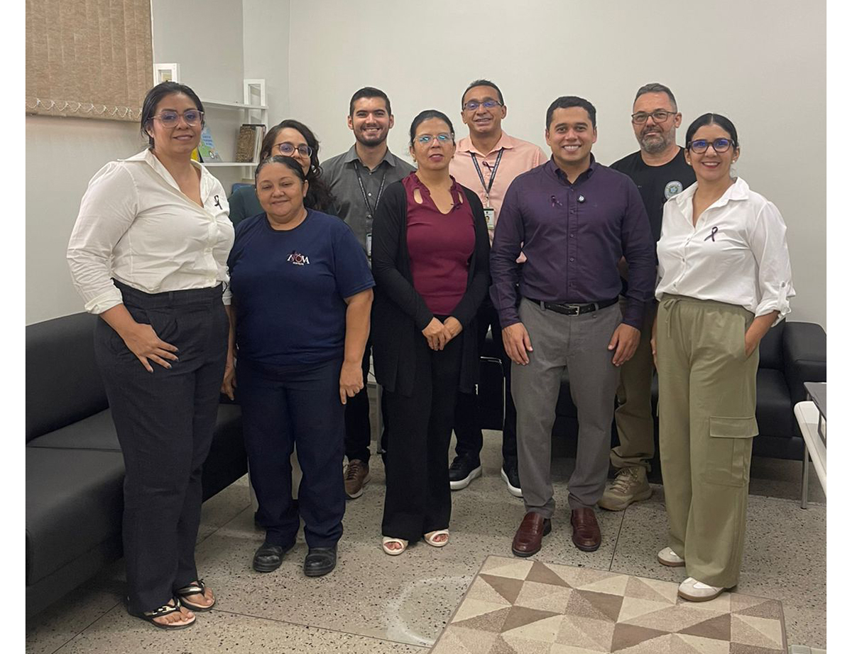 O médico perito Anisio Pinheiro, de camisa roxa, ministra palestra para servidores e estagiários da unidade, que o escutam atentamente sentados em sofás pretos. O grupo também posa para uma foto oficial, onde é possível observar que vários integrantes utilizam o laço lilás na roupa, símbolo da campanha de conscientização. O evento ocorre em uma sala com paredes brancas, estante de livros ao fundo e um tapete com formas geométricas no centro