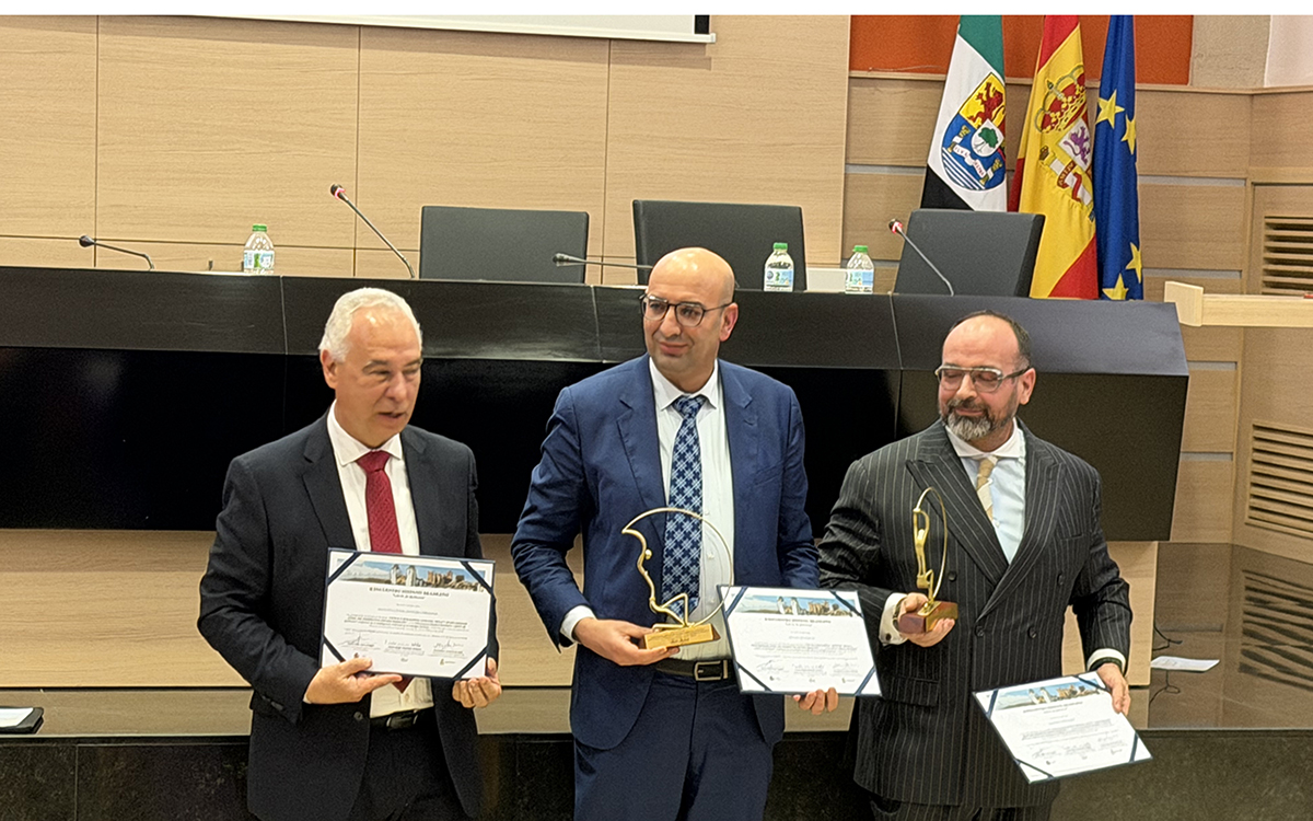 Três homens de terno posam à frente de uma bancada preta em um auditório, segurando certificados e troféus dourados de formato circular. O homem à esquerda usa gravata vermelha, o do centro é calvo e usa gravata azul quadriculada, e o da direita veste um terno risca-de-giz com gravata bege. Ao fundo, as bandeiras da Extremadura, Espanha e União Europeia completam o cenário institucional.