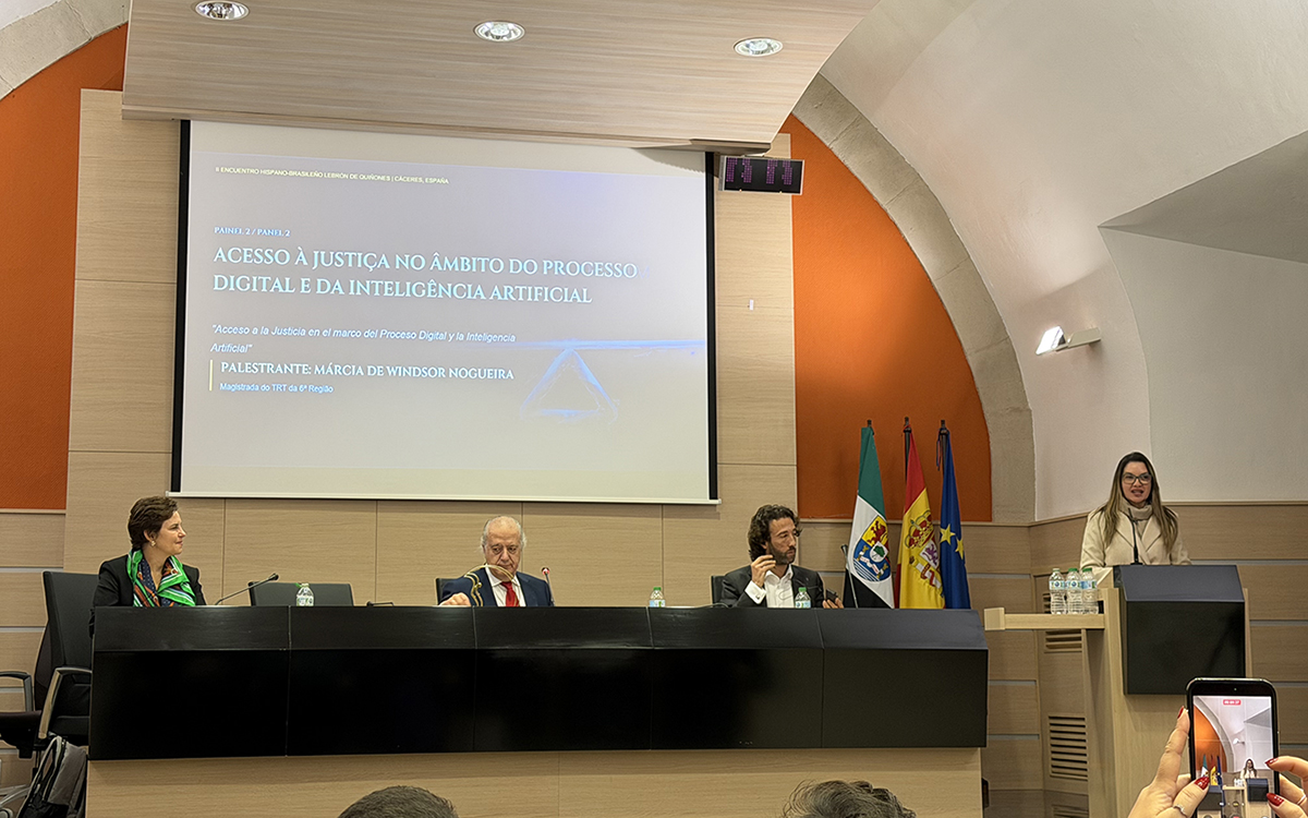 A magistrada Márcia de Windsor Nogueira, de cabelos longos e blazer bege, discursa em um púlpito durante o Painel 2 sobre acesso à justiça e inteligência artificial. No centro, uma mesa diretora acomoda três integrantes que a observam, tendo ao fundo um grande telão com o título da palestra e as bandeiras institucionais. Em primeiro plano, no canto inferior direito, as mãos de uma pessoa seguram um celular registrando o evento.