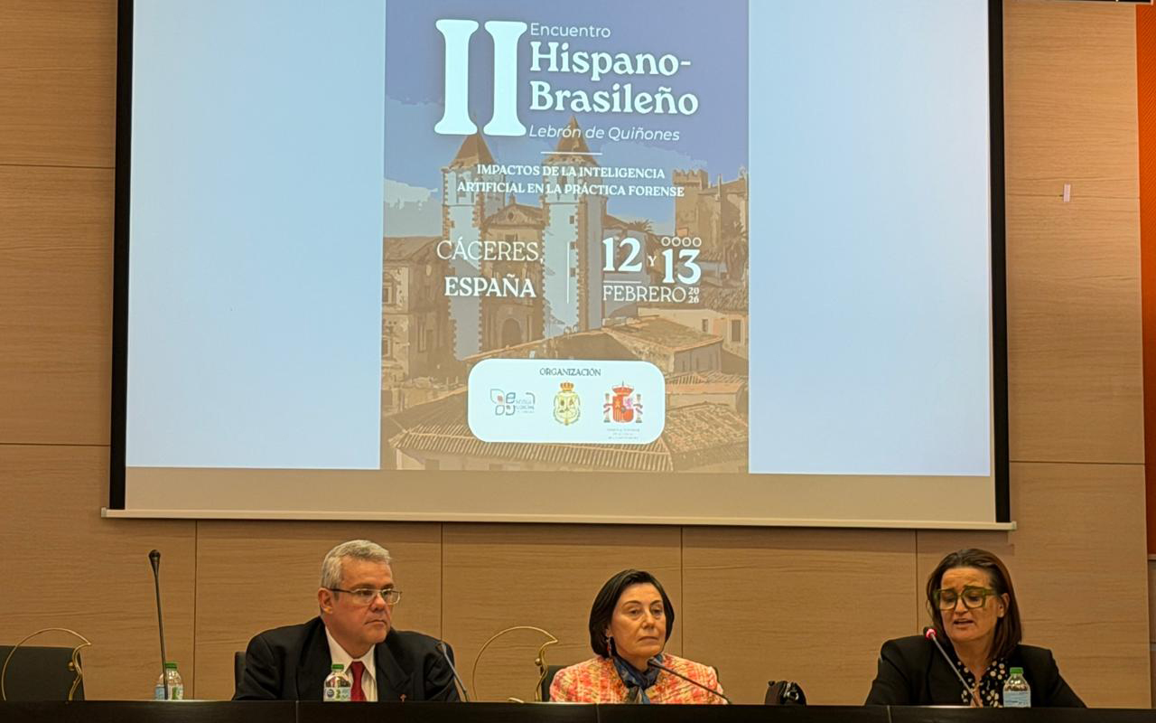 Três integrantes do encontro, dois homens e uma mulher, compõem a mesa de debates diante de uma bancada preta. Ao fundo, um telão exibe o cartaz do "II Encuentro Hispano-Brasileño Lebrón de Quiñones", destacando o tema sobre Inteligência Artificial, a cidade de Cáceres e as datas de 12 e 13 de fevereiro de 2026. O ambiente mantém o padrão institucional com painéis de madeira clara e iluminação focal.