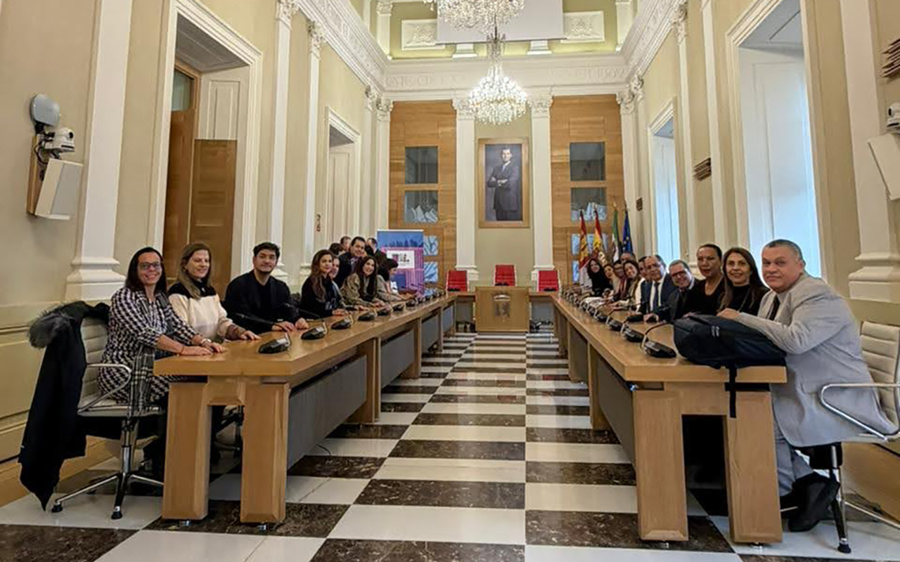 Foto que apresenta os juristas sentados ao longo de uma grande mesa retangular em um salão clássico com teto alto e lustres de cristal. Homens e mulheres vestem trajes formais e sorriem para a câmera em ambiente institucional iluminado.