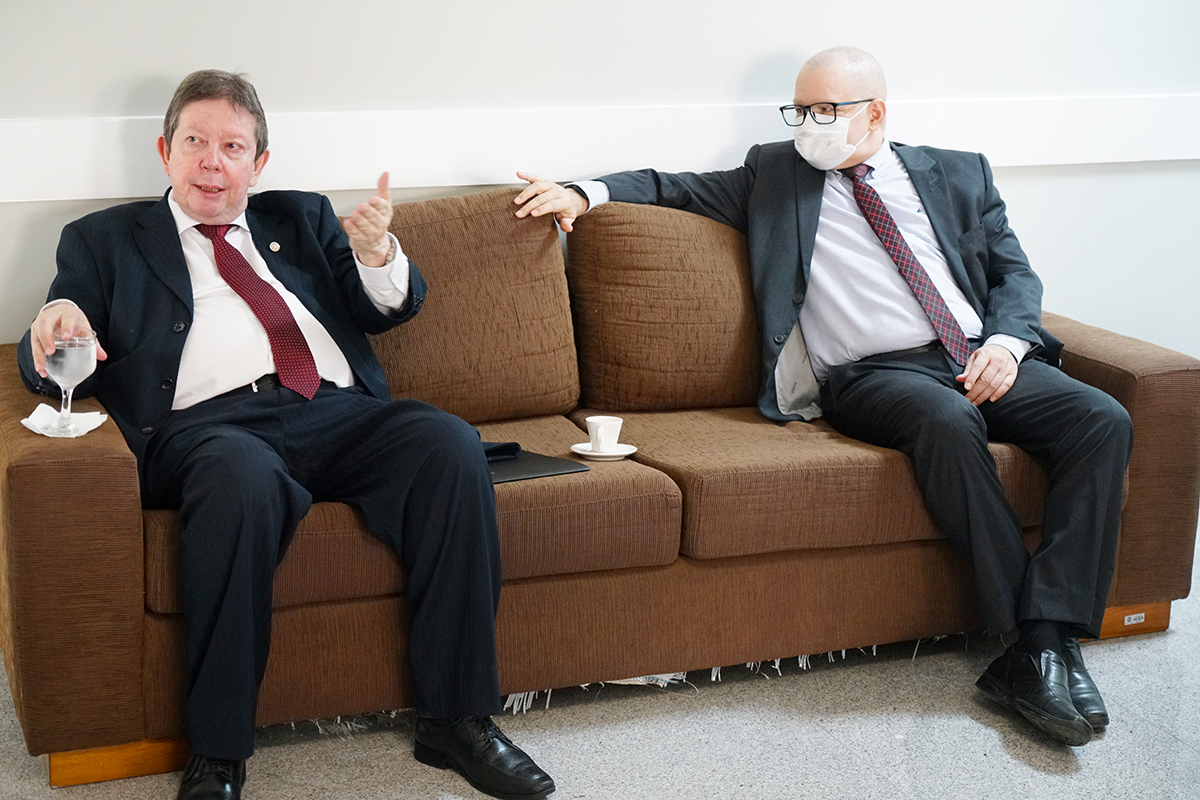 Dois homens conversam sentados em um sofá marrom. O homem à esquerda gesticula enquanto fala, segurando uma taça de água. O homem à direita, usando óculos e máscara de proteção branca, ouve atentamente. Ambos vestem ternos escuros e camisas claras com gravata.