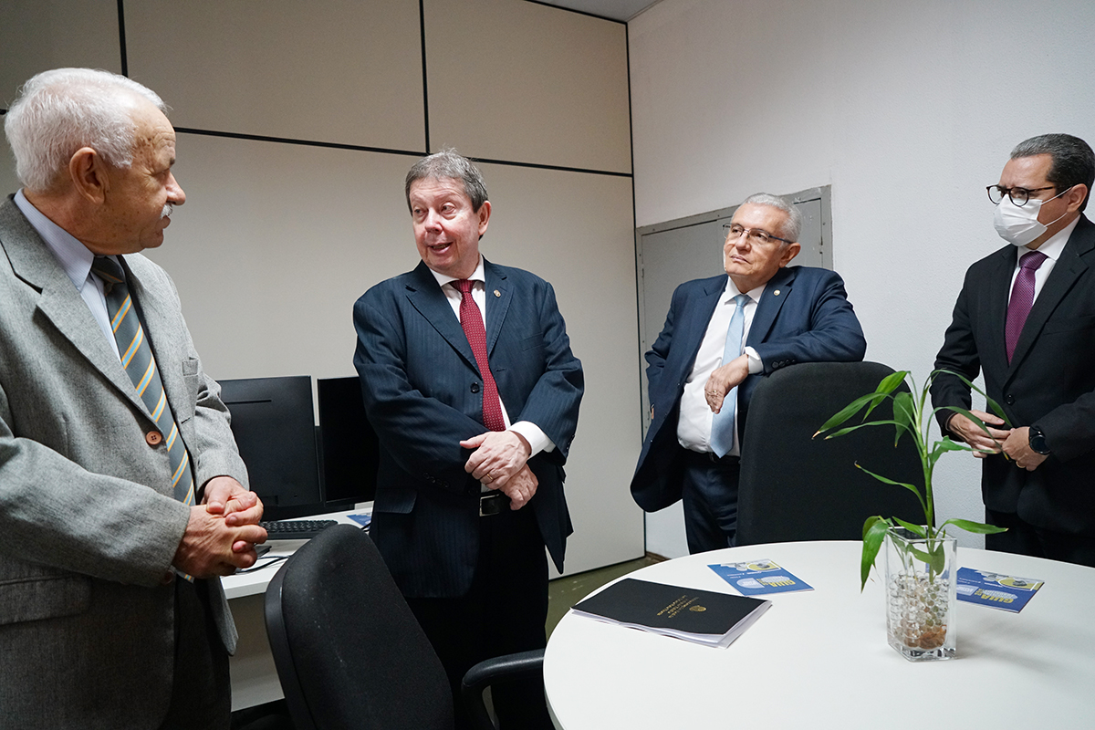 Quatro homens em trajes formais conversam em pé ao redor de uma mesa redonda branca com documentos e um pequeno vaso de planta. O ambiente possui divisórias de escritório claras. Três homens observam o que está ao centro, que fala e gesticula levemente, mantendo um tom de diálogo.
