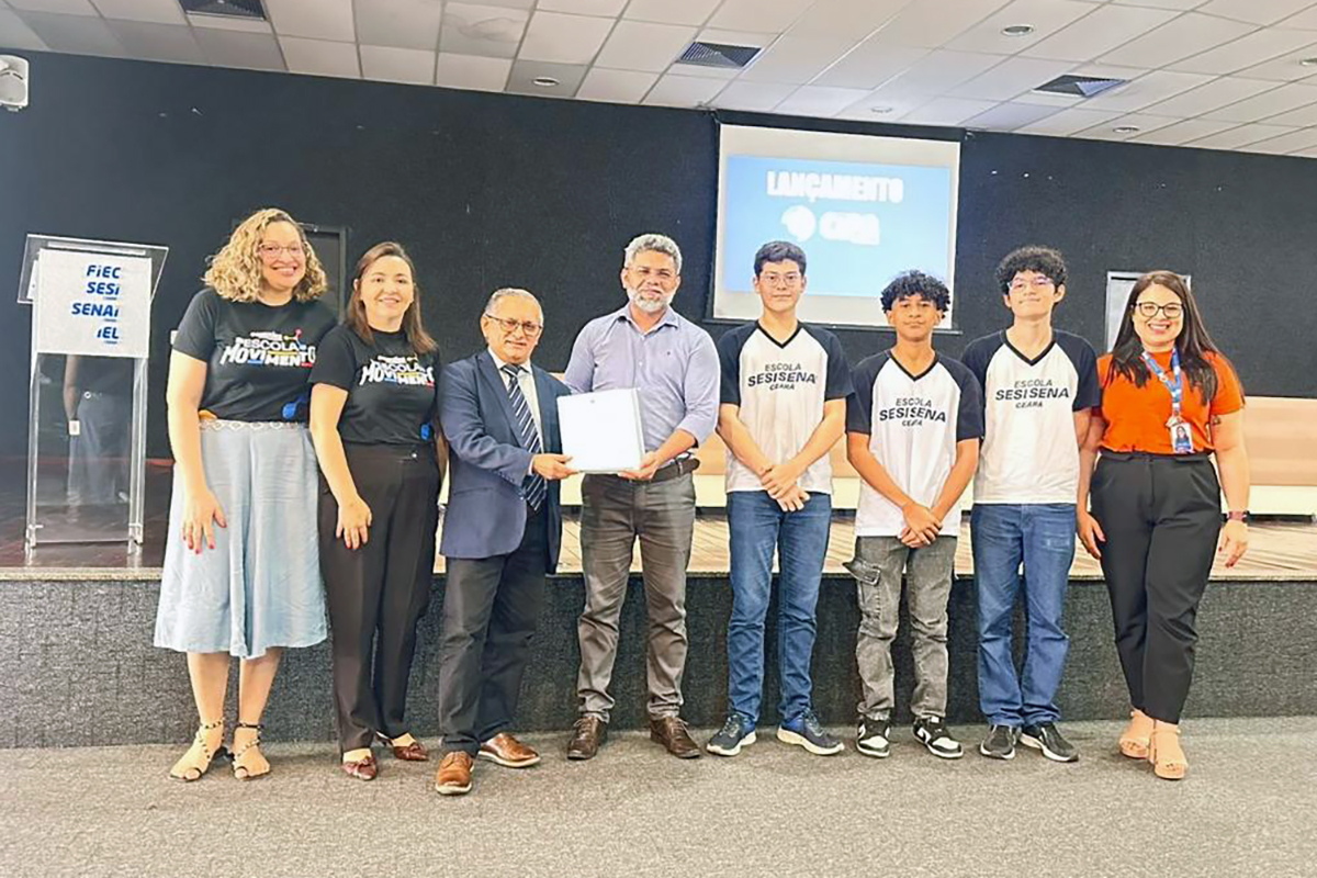 Oito pessoas posam lado a lado em um auditório: à esquerda, duas mulheres e dois homens em trajes casuais e formais; ao centro, três jovens com uniforme branco e azul da "Escola SESI SENAI"; à direita, uma mulher de blusa laranja. Os dois homens centrais seguram juntos um documento branco, com um telão ao fundo exibindo a palavra "LANÇAMENTO".