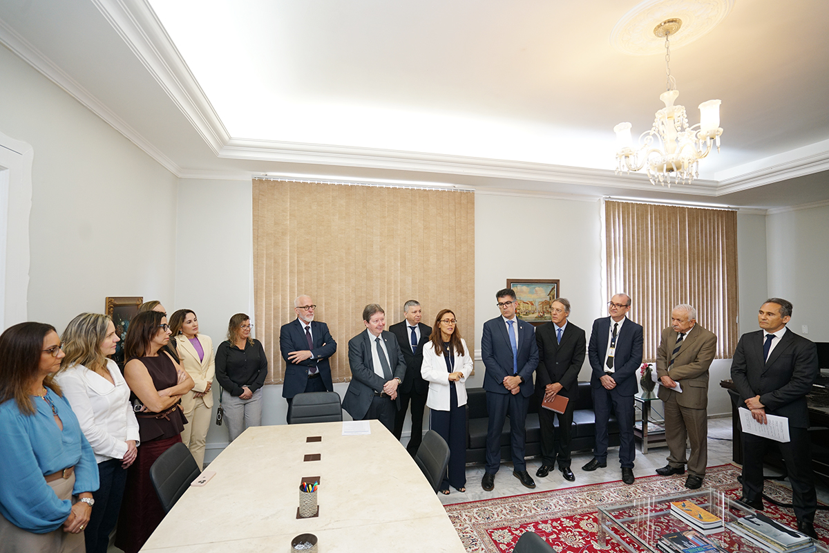 Grupo de aproximadamente quinze pessoas, homens e mulheres em trajes formais, posicionado em pé dentro de uma sala de reuniões. Eles estão dispostos em semicírculo sobre um tapete ornamentado, ouvindo uma mulher que fala ao centro. O ambiente possui tons claros, cortinas bege e um lustre de cristal.