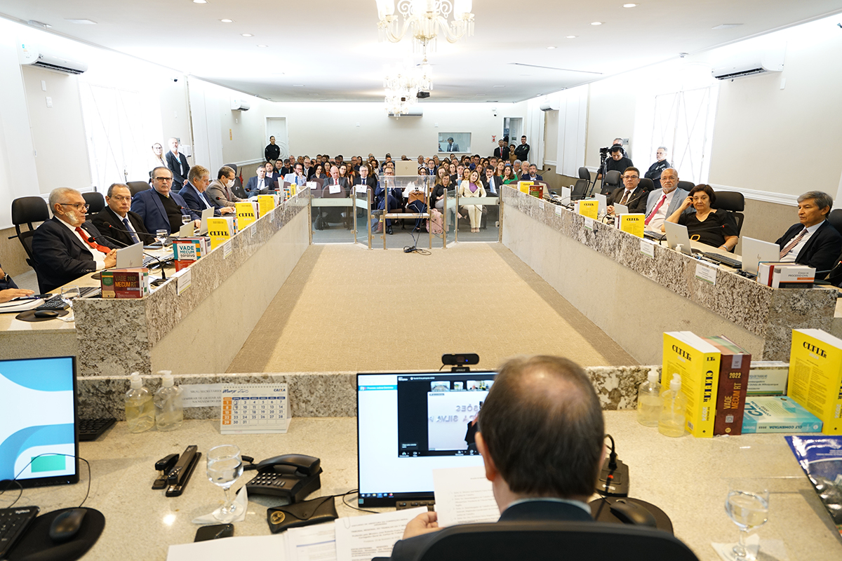 Amplo plenário com mesas em formato de "U" ocupadas por diversas autoridades de terno. Ao fundo, uma plateia assiste à reunião em uma sala bem iluminada por lustres clássicos. Em primeiro plano, vê-se a bancada com um monitor, vades mécuns coloridos e a cabeça de um homem de costas.