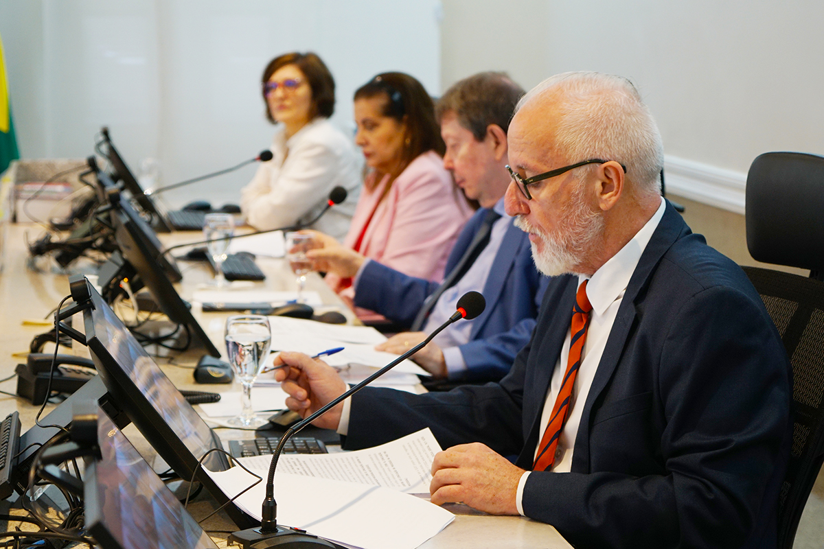 Close lateral da mesa diretora sob luz clara. Em primeiro plano, um homem calvo de óculos e barba branca lê papéis atentamente. Ao fundo, de forma levemente desfocada, aparecem os outros integrantes da mesa e equipamentos de informática, destacando o ambiente de trabalho oficial.