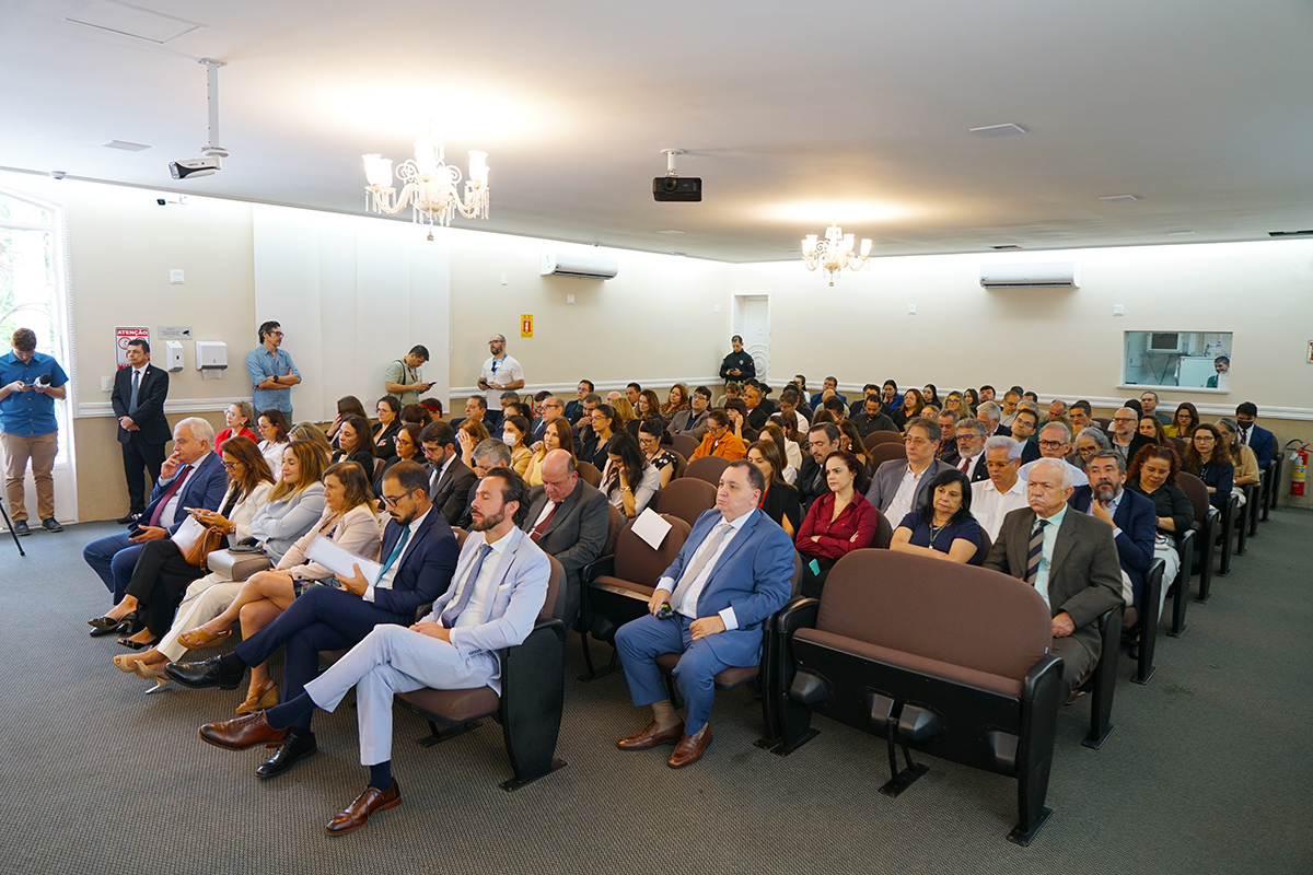 Visão ampla do auditório ocupado por dezenas de pessoas sentadas em poltronas marrons. O público, em trajes formais e casuais, assiste à cerimônia. O teto branco possui lustres clássicos e aparelhos de ar-condicionado, enquanto as paredes laterais têm janelas e portas brancas.