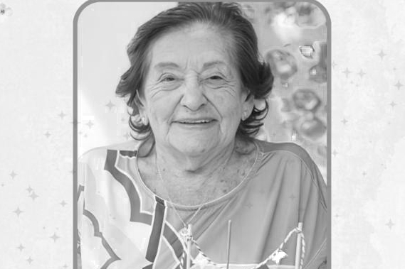 Retrato em preto e branco de uma mulher idosa, sorridente, de pele clara e cabelos curtos e ondulados. Ela usa uma blusa com estampas geométricas e um colar fino com pingente. A imagem possui uma moldura cinza de cantos arredondados, sobre um fundo claro com ilustrações sutis de estrelas e brilhos.