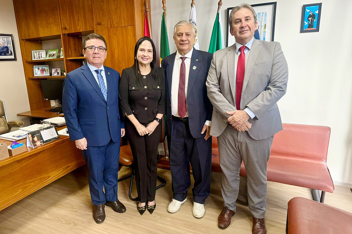 Quatro pessoas — três homens e uma mulher — posam em pé dentro de um escritório. Os homens vestem ternos e gravatas, enquanto a mulher usa um conjunto preto com botões frontais. Eles estão alinhados à frente de uma mesa de madeira e bandeiras oficiais, em um ambiente com piso de madeira clara e sofás de couro ao fundo.