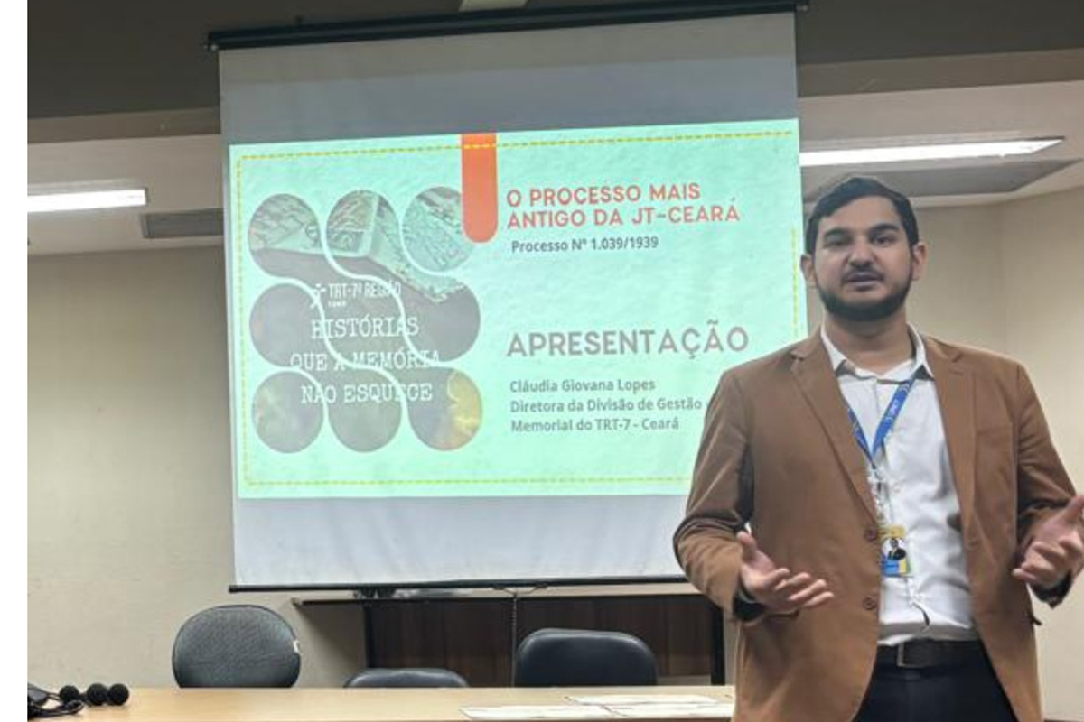 Um homem jovem, com barba e vestindo blazer caramelo, realiza uma palestra à frente de uma tela de projeção. O slide exibe o título "O processo mais antigo da JT-Ceará" e menciona Cláudia Giovana Lopes, diretora do Memorial do TRT-7. Ele gesticula com as mãos em um auditório com mesa de madeira, cadeiras pretas e piso acarpetado.