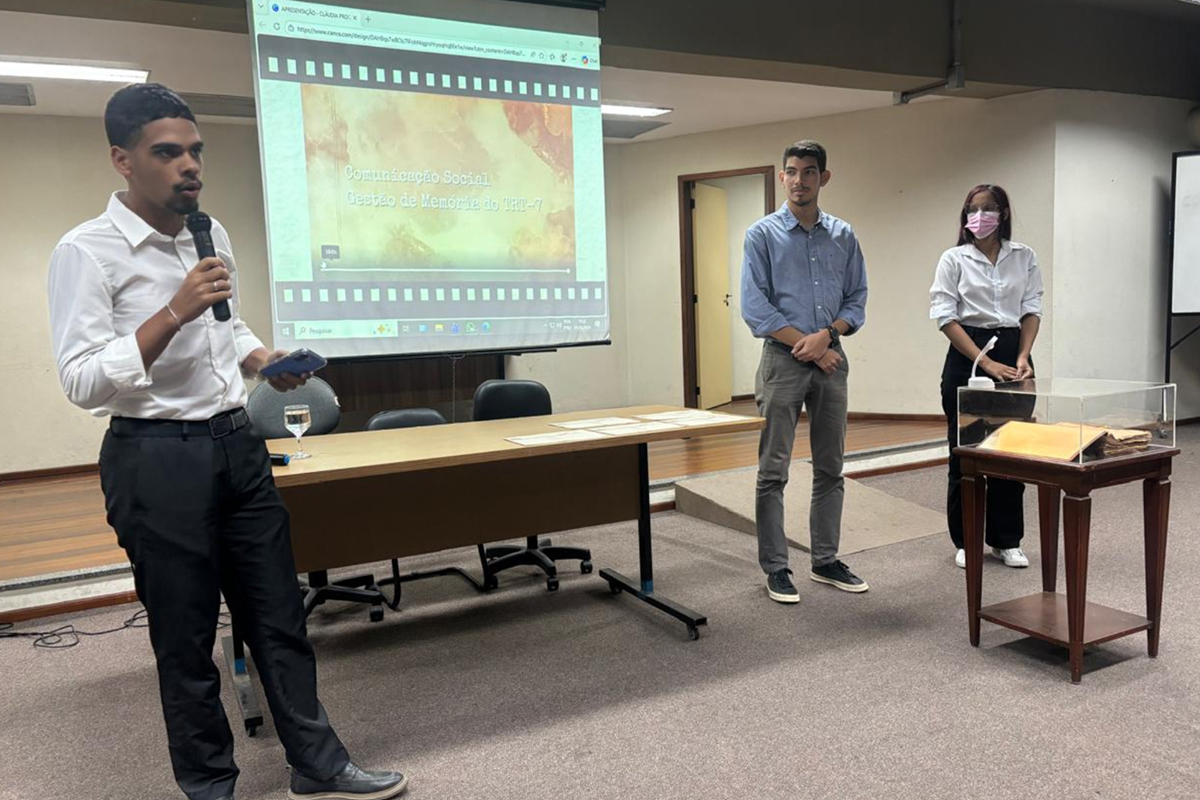 Um jovem de camisa branca e calça preta fala ao microfone diante de uma tela que projeta o título "Comunicação Social - Gestão de Memória do TRT-7". Ao lado, dois outros jovens acompanham a apresentação em um auditório com mesa de madeira e piso acarpetado. No canto direito, destaca-se uma pequena mesa com uma redoma de acrílico, protegendo um documento antigo e amarelado.