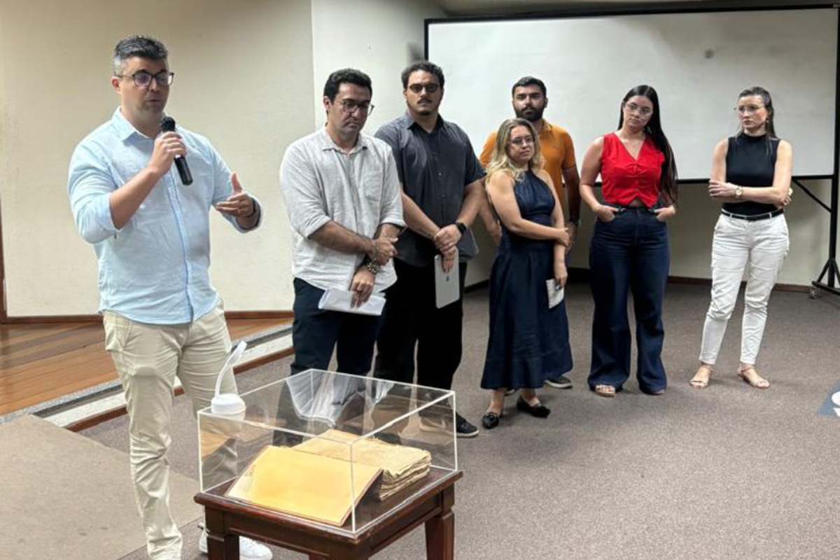 Um homem de camisa azul clara e óculos fala ao microfone, liderando um grupo de seis pessoas em um auditório com piso acarpetado. Em primeiro plano, uma mesa de madeira sustenta uma redoma de acrílico que protege um volumoso processo jurídico antigo, com páginas amareladas. O grupo observa atentamente a apresentação, posicionando-se à frente de uma tela de projeção branca ao fundo.