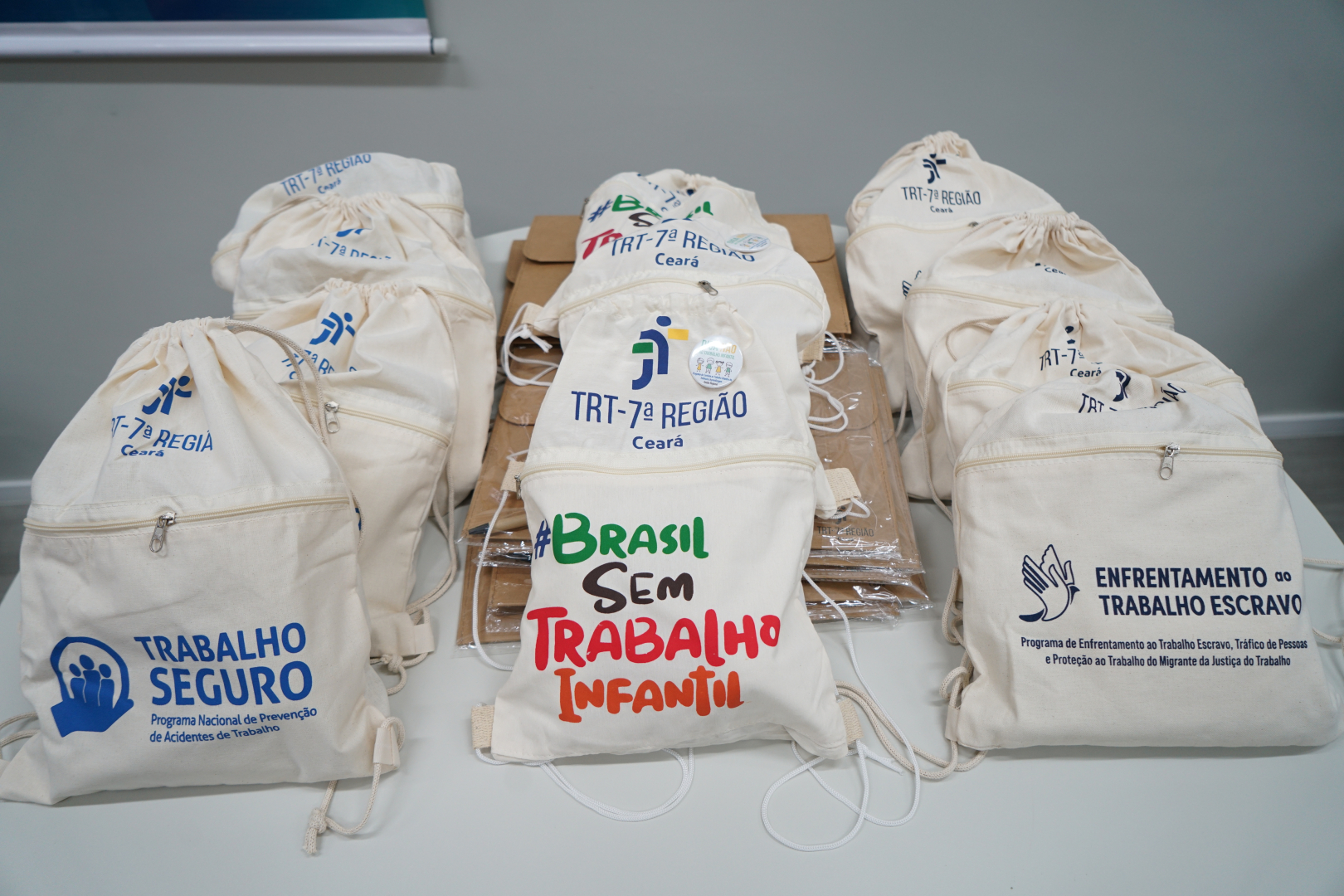 A imagem, em plano médio e levemente de cima para baixo, apresenta diversos mochilas tipo saco (ecobags) de cor crua, organizadas sobre uma mesa branca. As mochilas são feitas de tecido de algodão e possuem cordões brancos para fechamento e alças.  As mochilas estão divididas em três grupos principais, cada um com uma estampa diferente referente a programas do TRT-7ª Região (Ceará):  À esquerda: Mochilas com o logotipo azul do programa "Trabalho Seguro" (Programa Nacional de Prevenção de Acidentes de Trabalho).  Ao centro: Mochilas com a hashtag colorida "#BRASIL SEM TRABALHO INFANTIL". As palavras estão em verde, marrom e vermelho. Acima da frase, vê-se o logotipo do TRT-7 e um pequeno bóton colorido.  À direita: Mochilas com a ilustração de uma mão aberta em forma de pomba e o texto em azul escuro: "ENFRENTAMENTO ao TRABALHO ESCRAVO".  Ao fundo, entre as pilhas de mochilas, notam-se envelopes pardos empilhados. A iluminação é clara e uniforme, sugerindo um ambiente institucional.